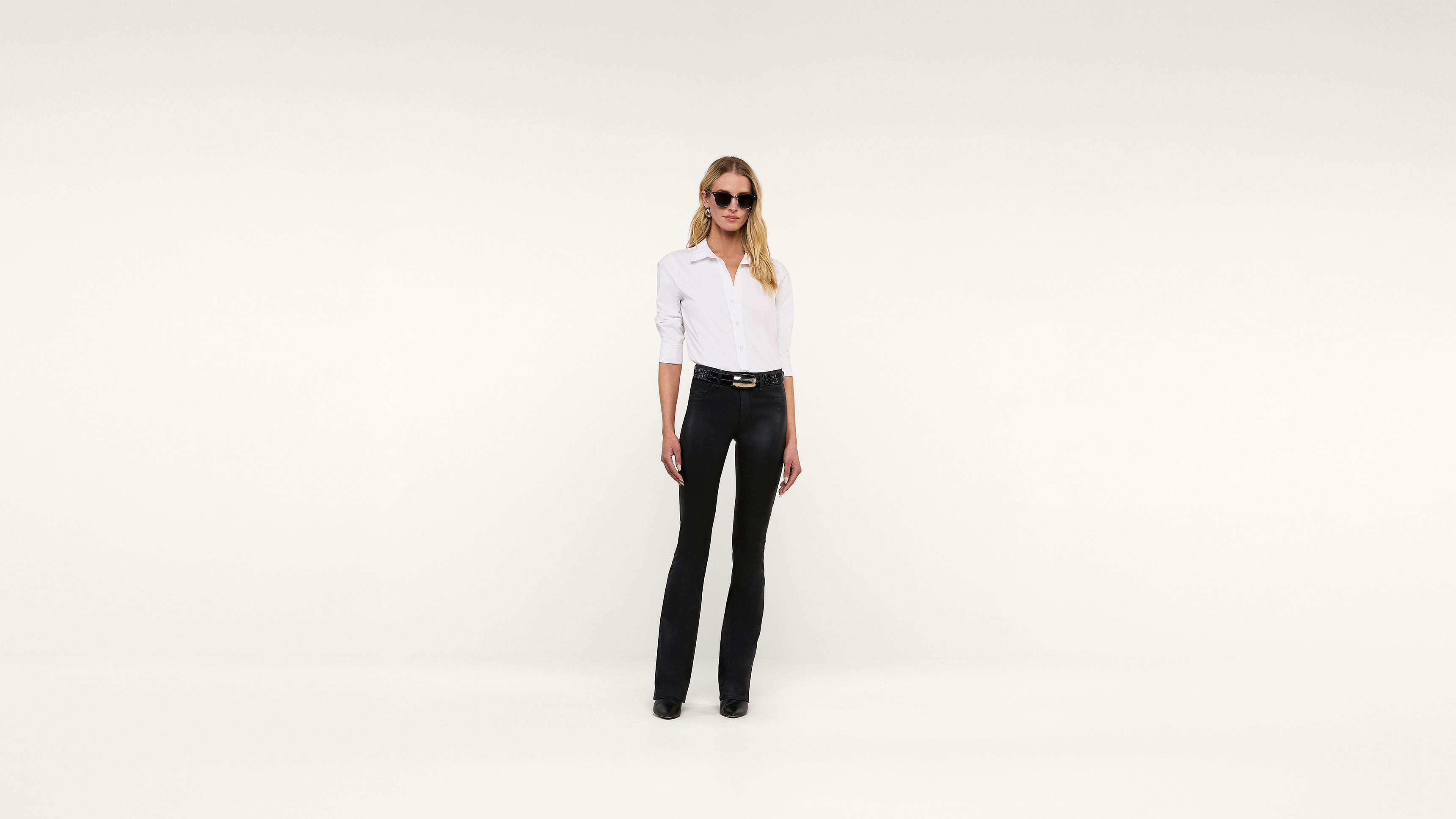 A woman wearing sunglasses, a white shirt, and black pants stands confidently against a plain white background, facing forward with her hands relaxed by her sides.