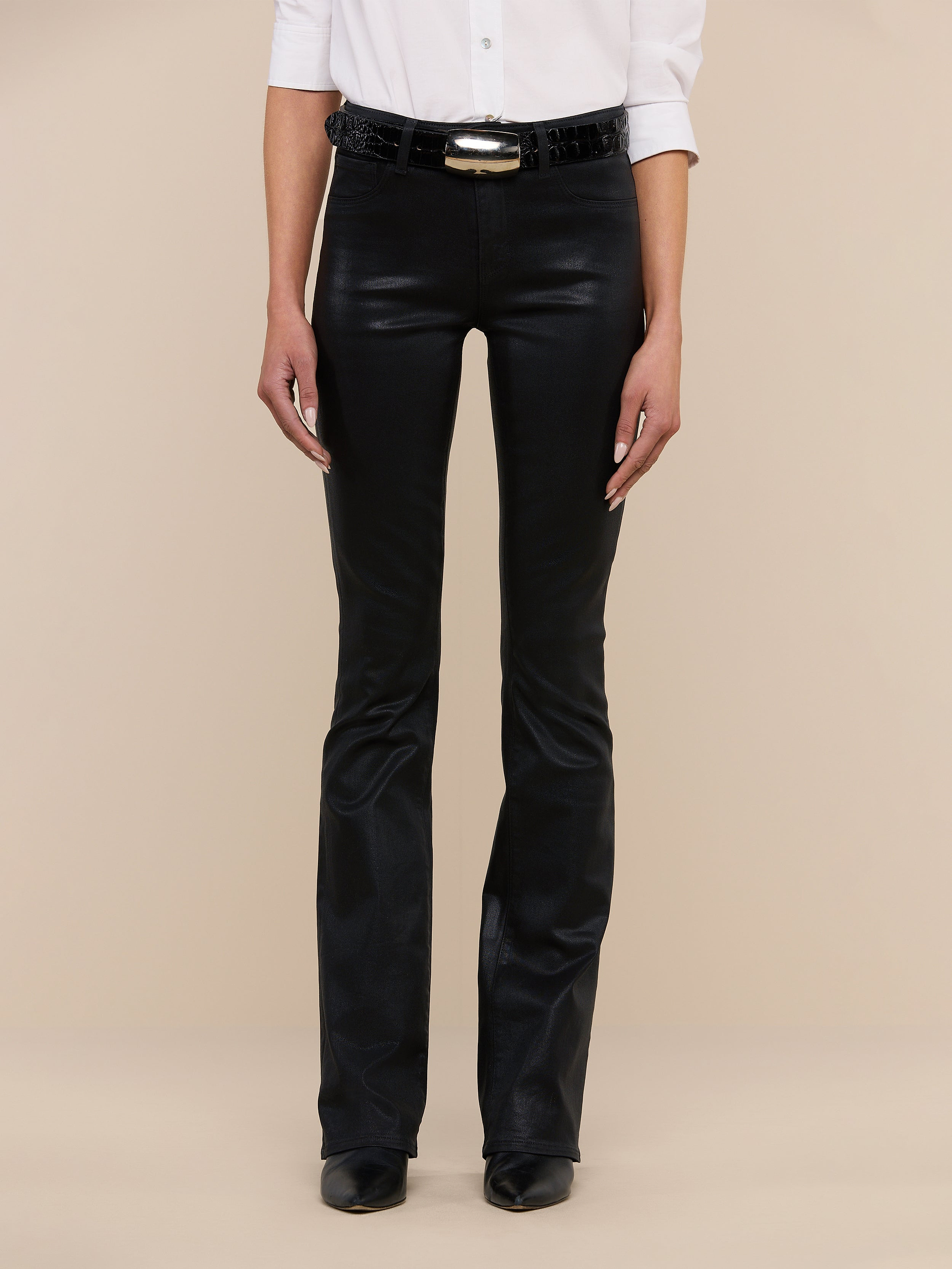 A person stands against a plain white background wearing the Selma Coated Bootcut Jean, styled with a white button-up shirt, a black belt, and black shoes. Their face is not visible.