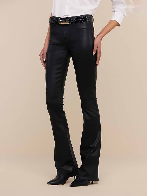 Wearing the Selma Coated Bootcut Jean, a white long-sleeve shirt, a black belt, and black pointed-toe boots, the person stands against a plain white background.