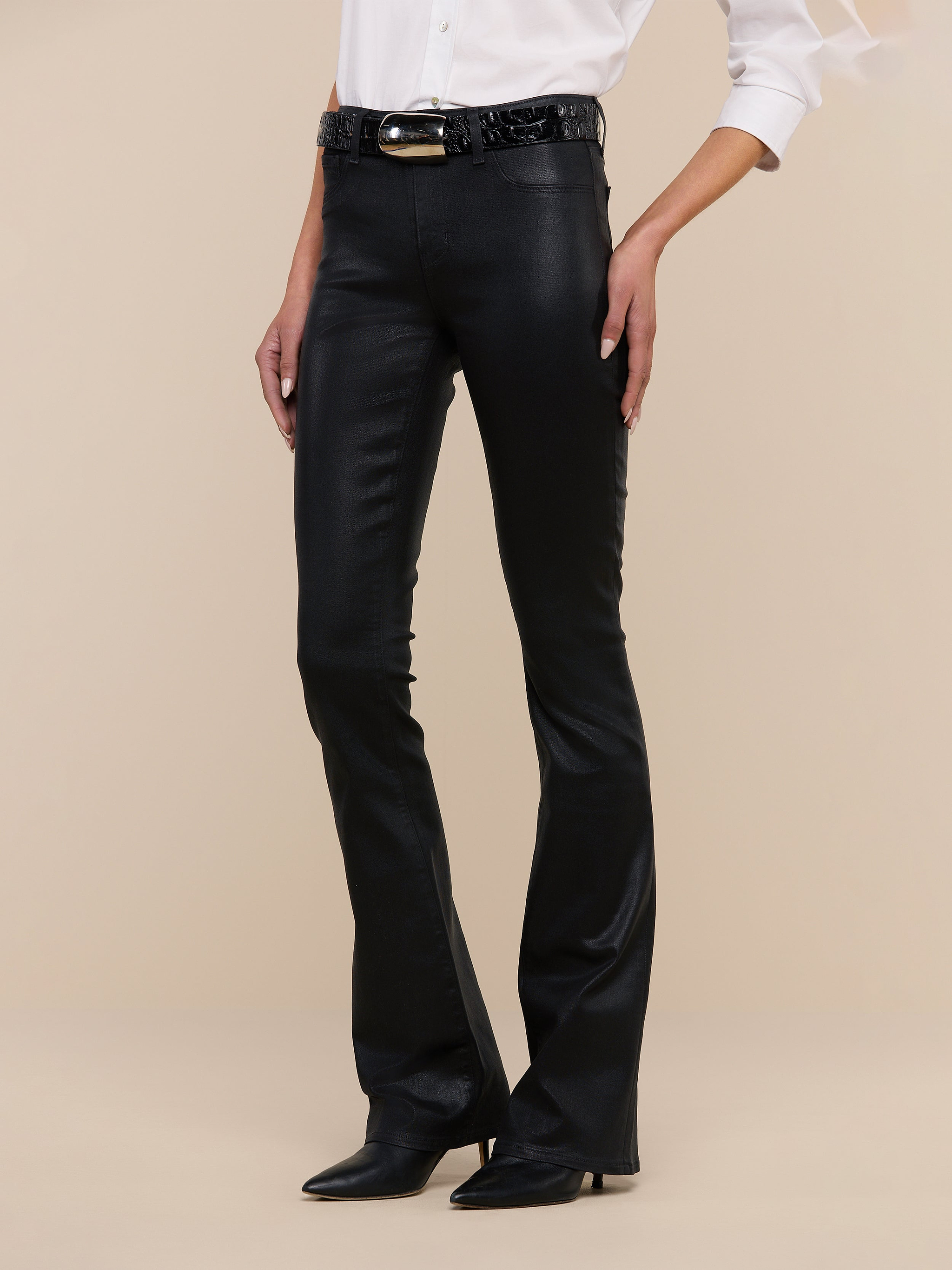 Wearing the Selma Coated Bootcut Jean, a white long-sleeve shirt, a black belt, and black pointed-toe boots, the person stands against a plain white background.
