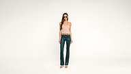 A woman wearing sunglasses, a beige sleeveless top, green leather pants, a tan belt, and open-toed heels stands against a plain white background.