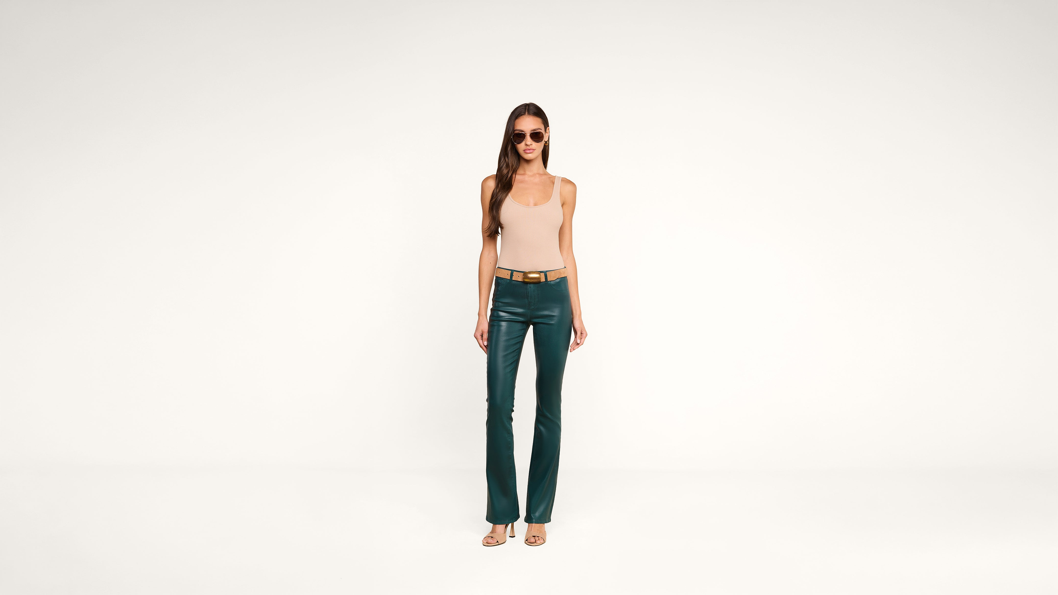 A woman wearing sunglasses, a beige sleeveless top, green leather pants, a tan belt, and open-toed heels stands against a plain white background.