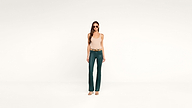 A woman wearing sunglasses, a beige tank top, green flare pants, and heels stands confidently against a plain white background.