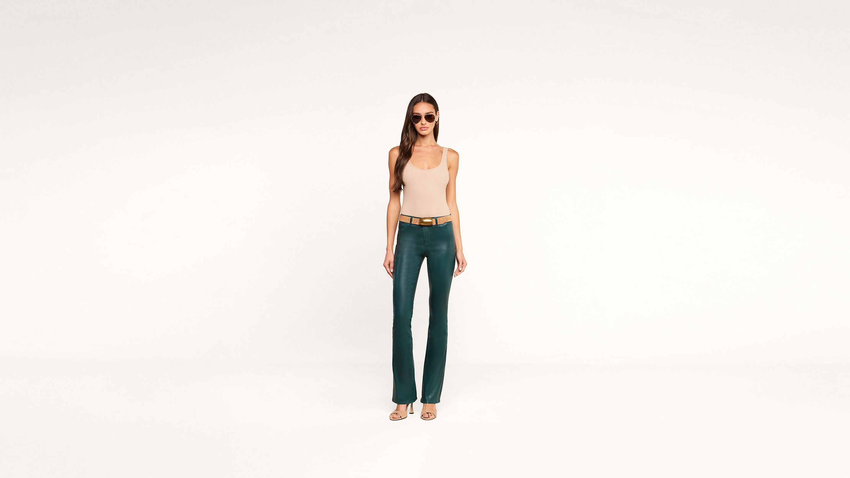 A woman wearing sunglasses, a beige tank top, green flare pants, and heels stands confidently against a plain white background.