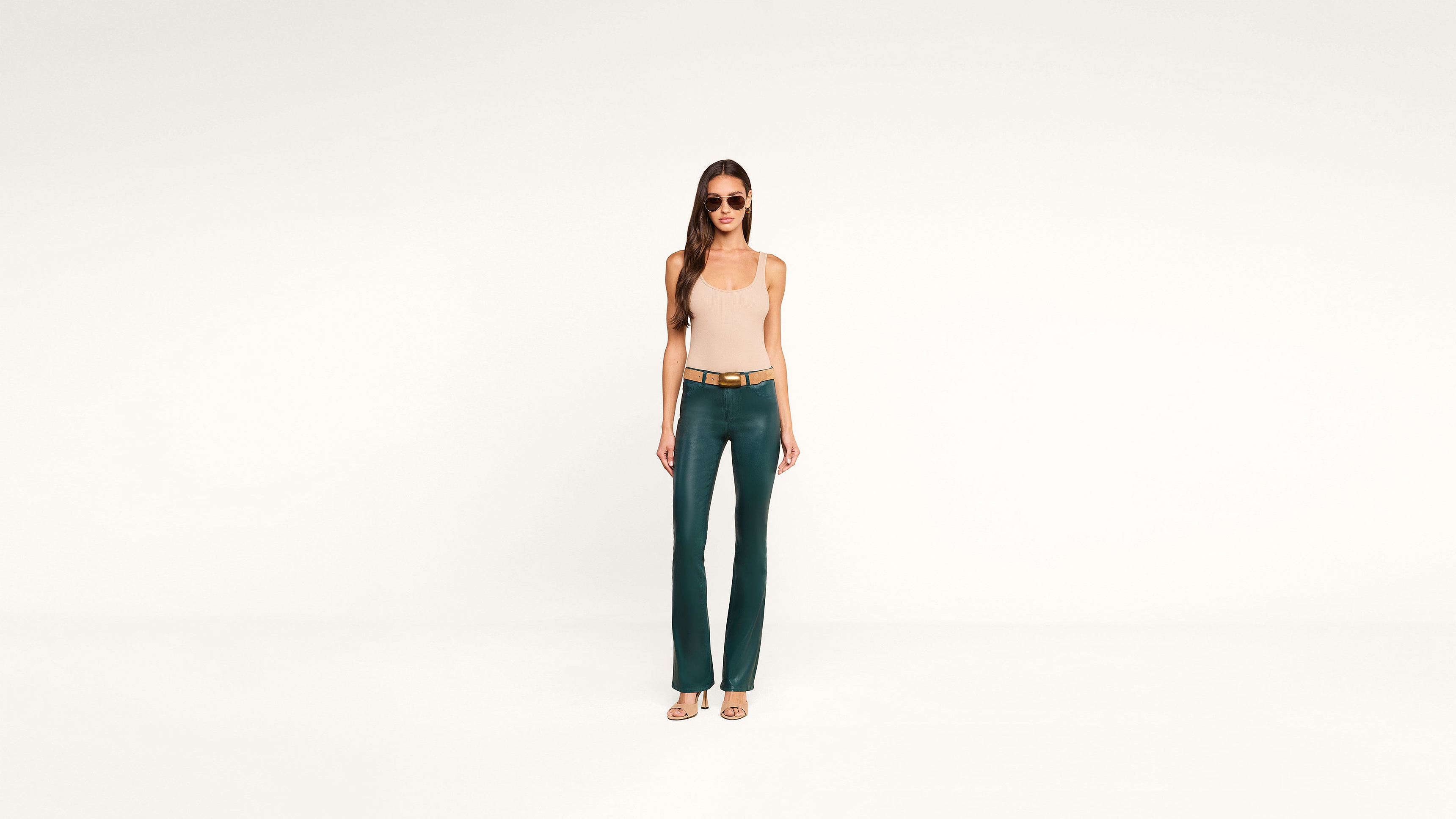 A woman wearing sunglasses, a beige tank top, green flare pants, and heels stands confidently against a plain white background.