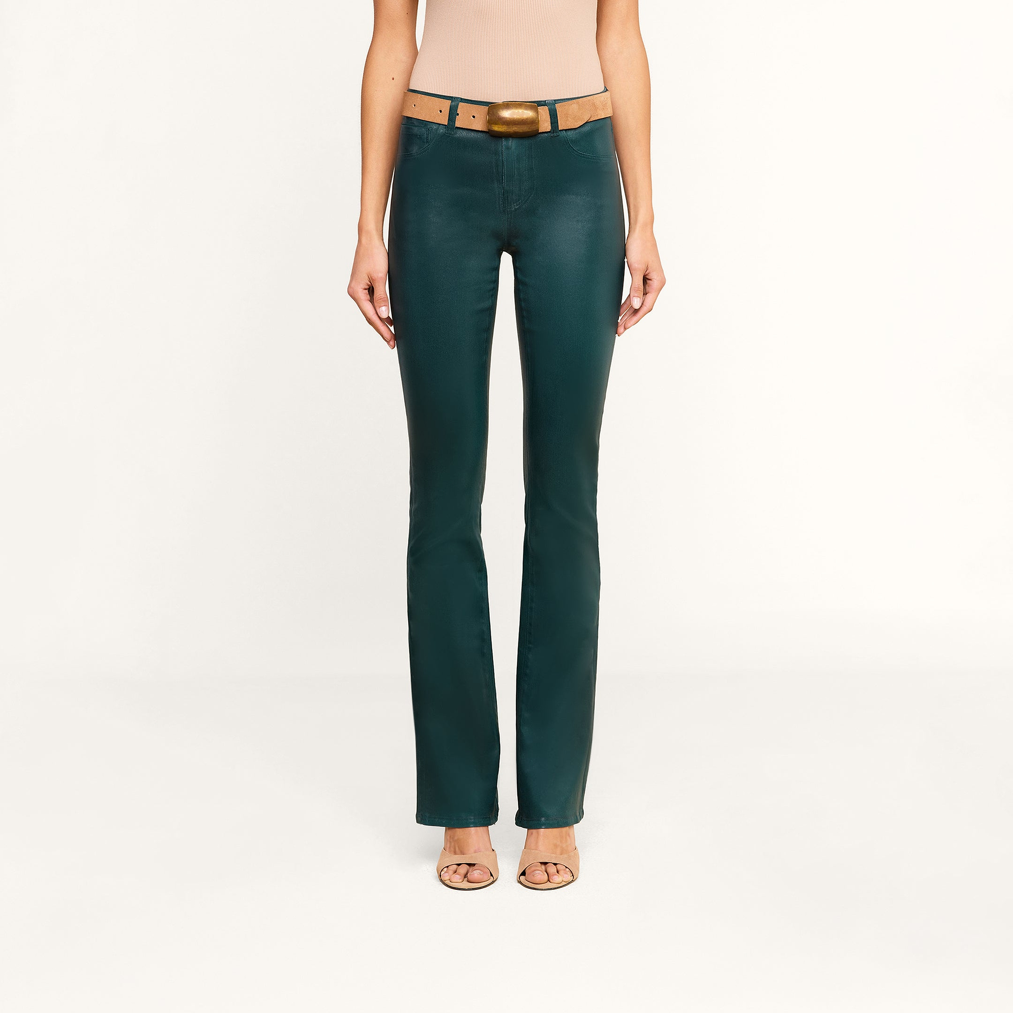 A woman models the Selma Coated Bootcut Jean in dark green, styled with a brown belt and nude heels, standing against a plain white background. Only her body from the shoulders down is shown.
