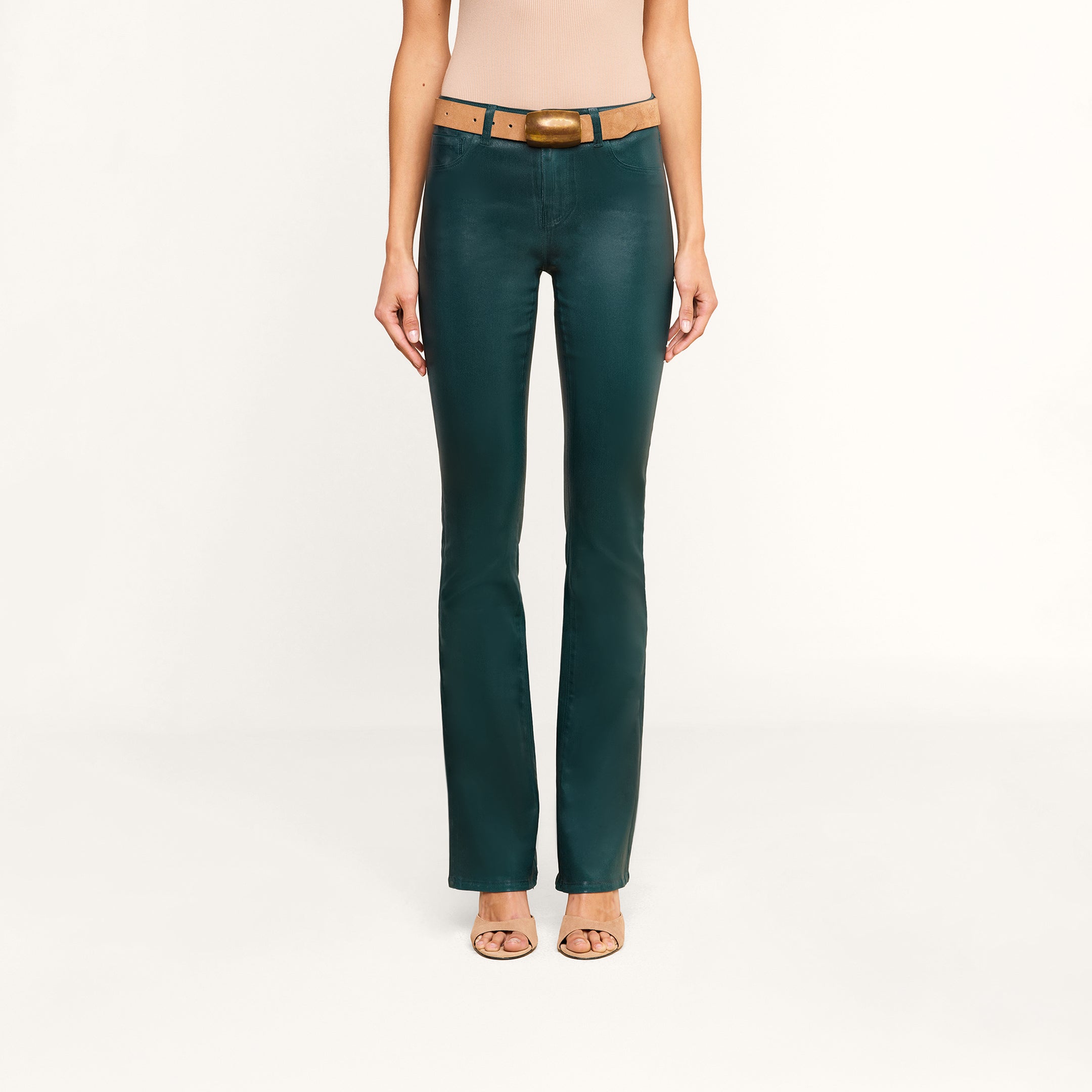 A woman models the Selma Coated Bootcut Jean in dark green, styled with a brown belt and nude heels, standing against a plain white background. Only her body from the shoulders down is shown.