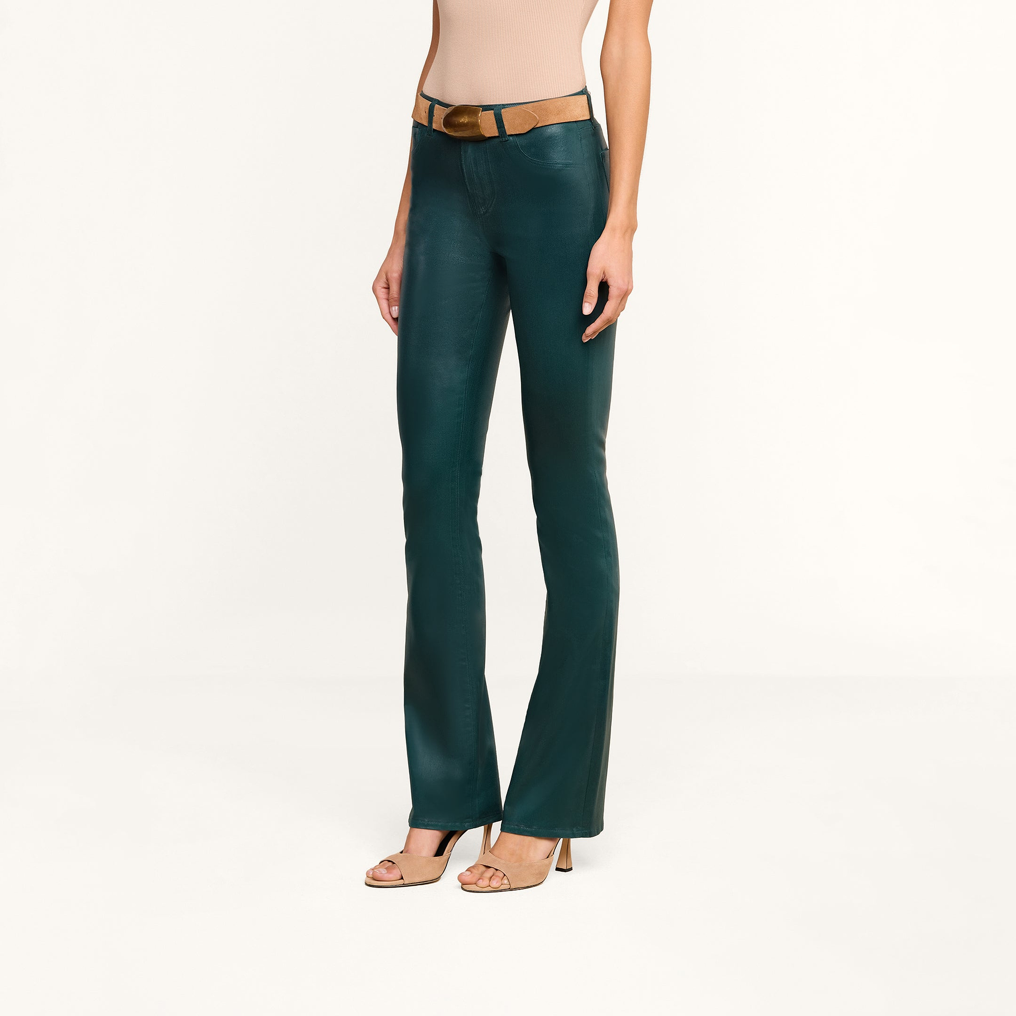 A person wearing the Selma Coated Bootcut Jean in dark green, a beige sleeveless top, a tan belt, and beige high-heeled sandals stands against a plain white background.