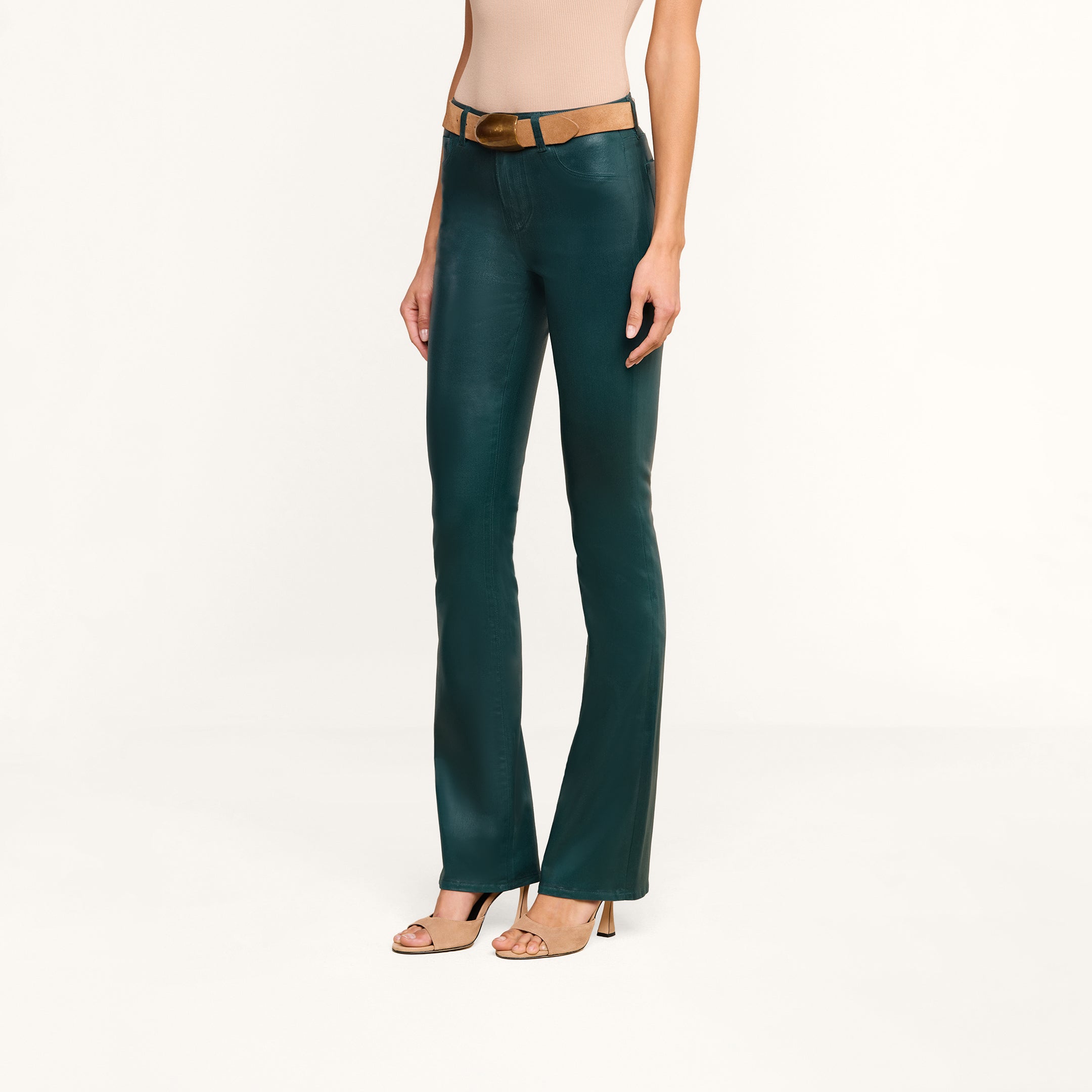 A person wearing the Selma Coated Bootcut Jean in dark green, a beige sleeveless top, a tan belt, and beige high-heeled sandals stands against a plain white background.
