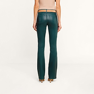 A woman stands facing away, wearing a beige sleeveless top and the Selma Coated Bootcut Jean in dark green with tan heels, against a plain white background.
