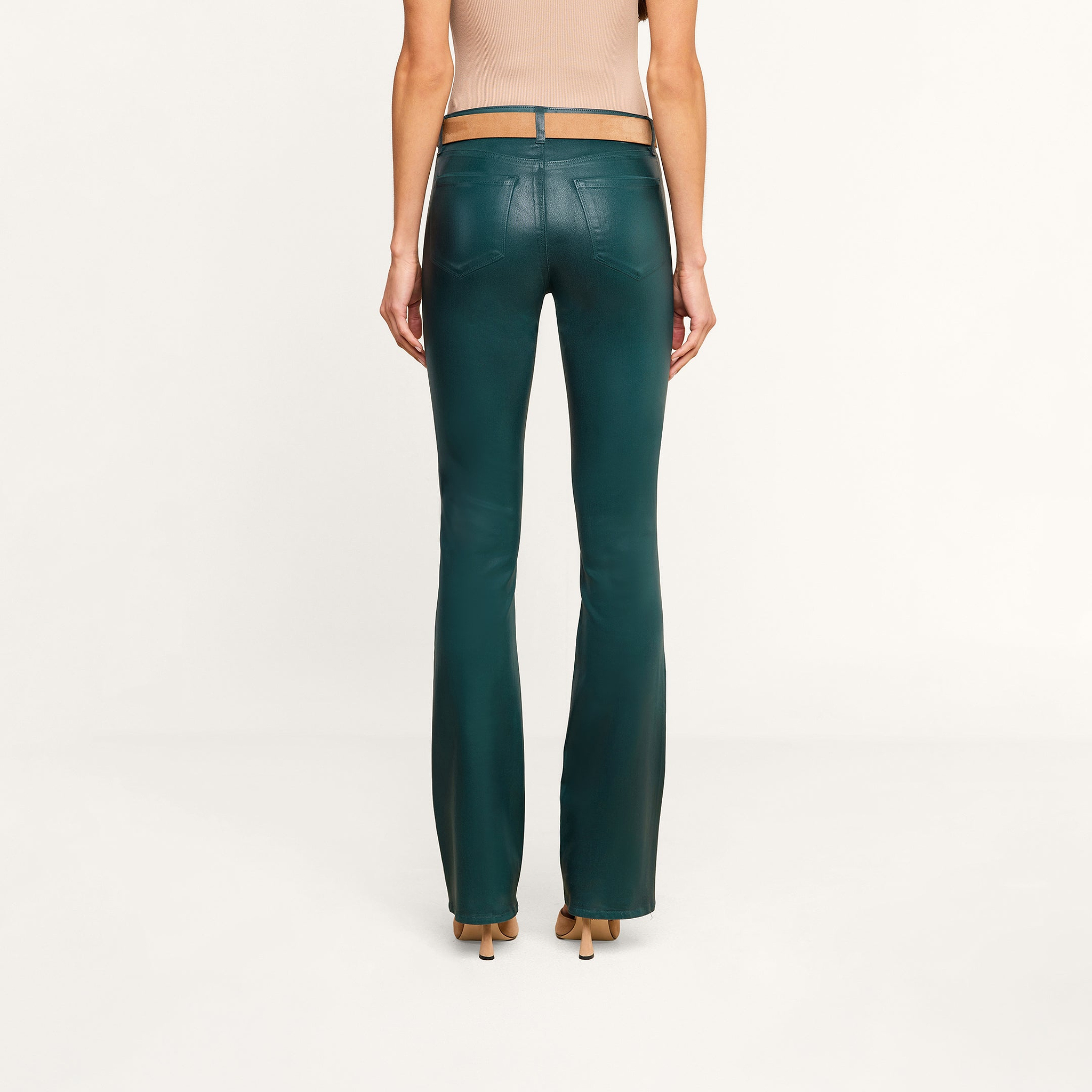 A woman stands facing away, wearing a beige sleeveless top and the Selma Coated Bootcut Jean in dark green with tan heels, against a plain white background.