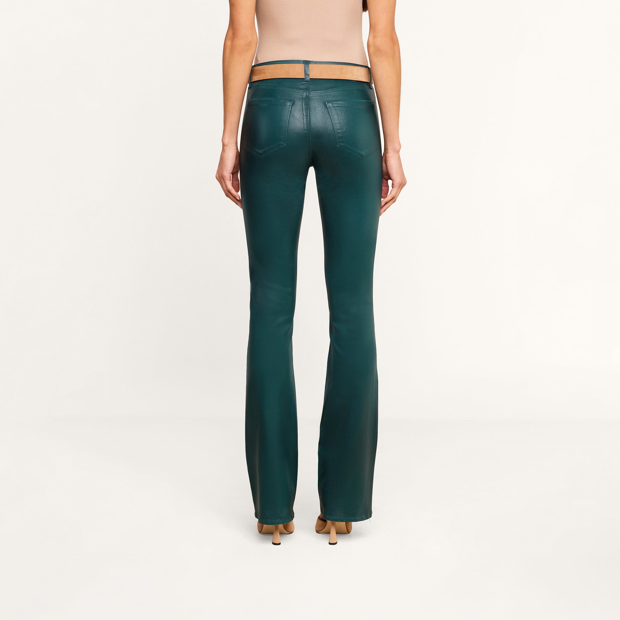 A woman stands facing away, wearing a beige sleeveless top and the Selma Coated Bootcut Jean in dark green with tan heels, against a plain white background.