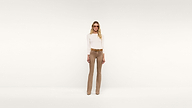 A person with long blonde hair wearing sunglasses, a white long-sleeve top, tan pants, a brown belt, and tan shoes stands stylishly against a plain white background.