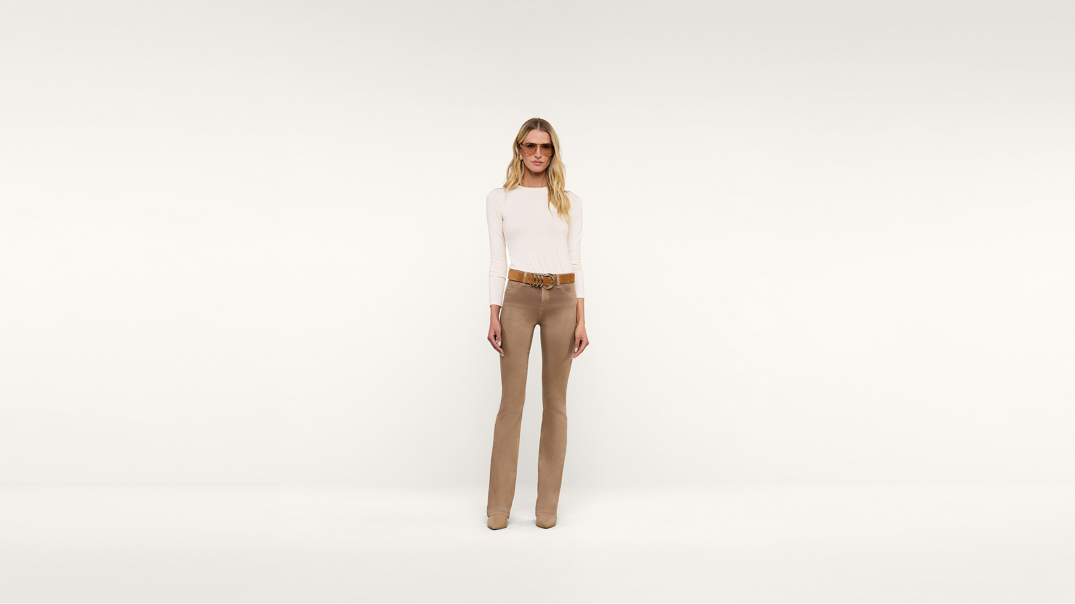 A person with long blonde hair wearing sunglasses, a white long-sleeve top, tan pants, a brown belt, and tan shoes stands stylishly against a plain white background.
