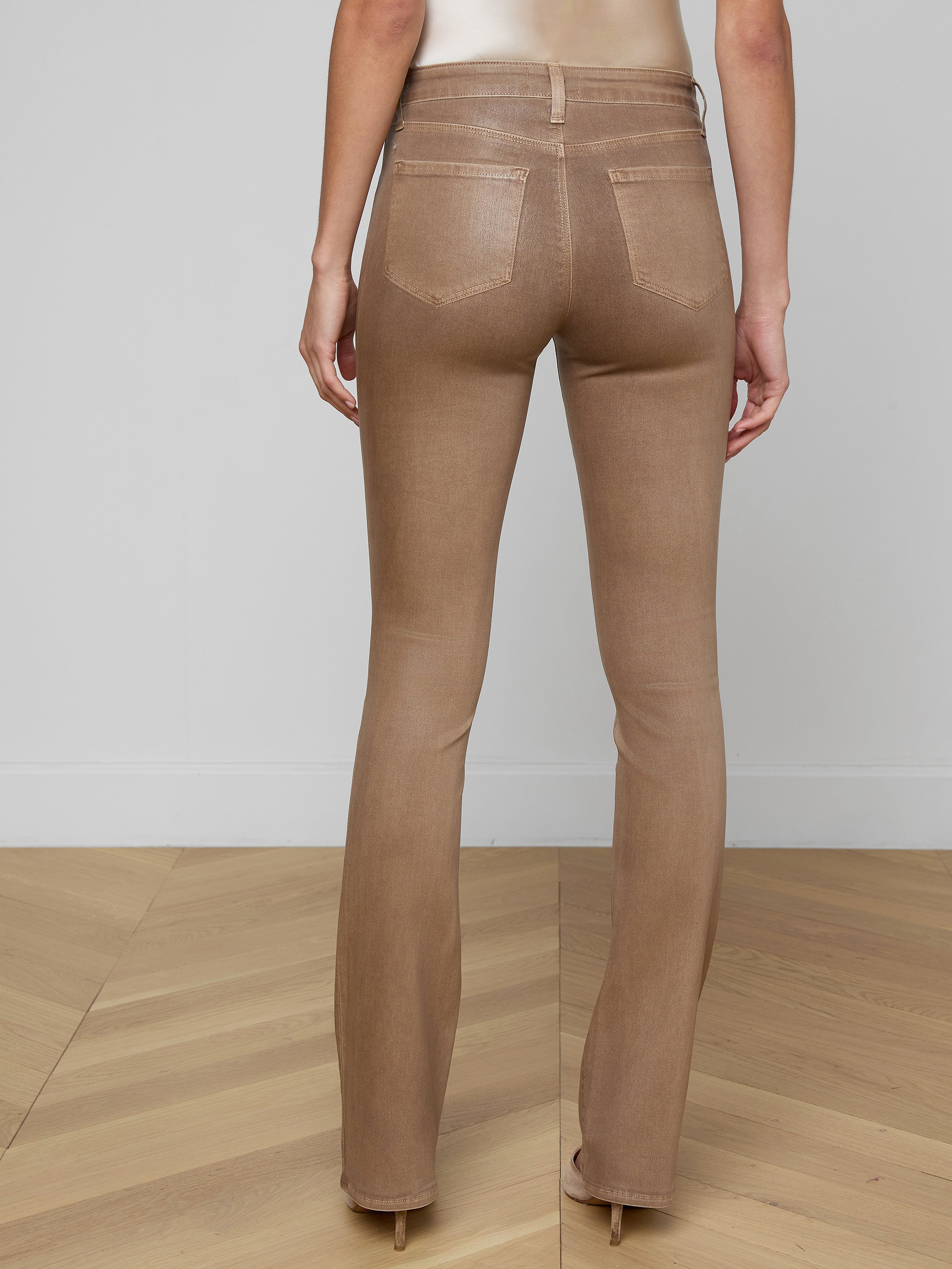 A person is shown from the back, lower half only, against a plain white background, wearing the Selma Coated Bootcut Jean in light brown, styled with a white long-sleeve top and a brown belt.