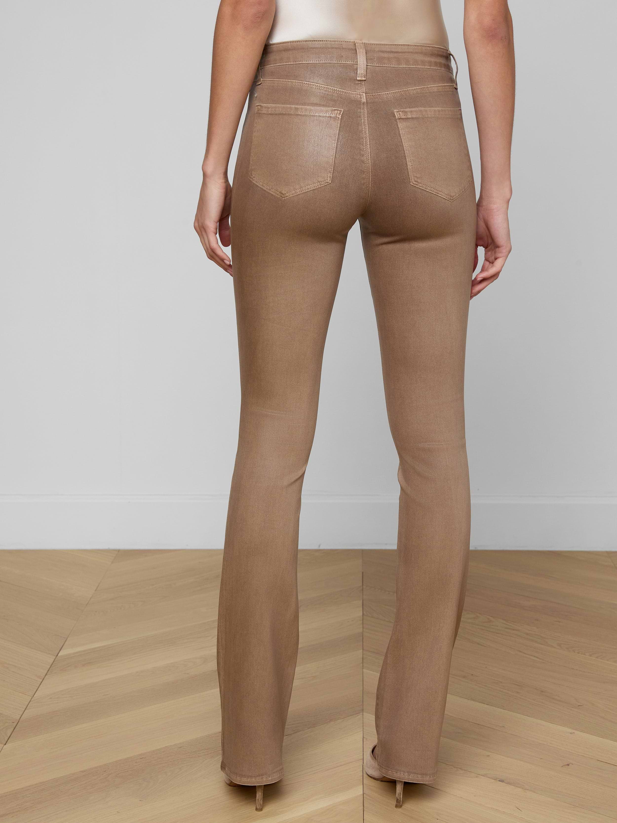 A person is shown from the back, lower half only, against a plain white background, wearing the Selma Coated Bootcut Jean in light brown, styled with a white long-sleeve top and a brown belt.