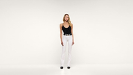 A woman stands against a plain white background wearing a black tank top, white pants, a black belt, and black shoes. She has blonde hair and a neutral expression, with her arms relaxed by her sides.