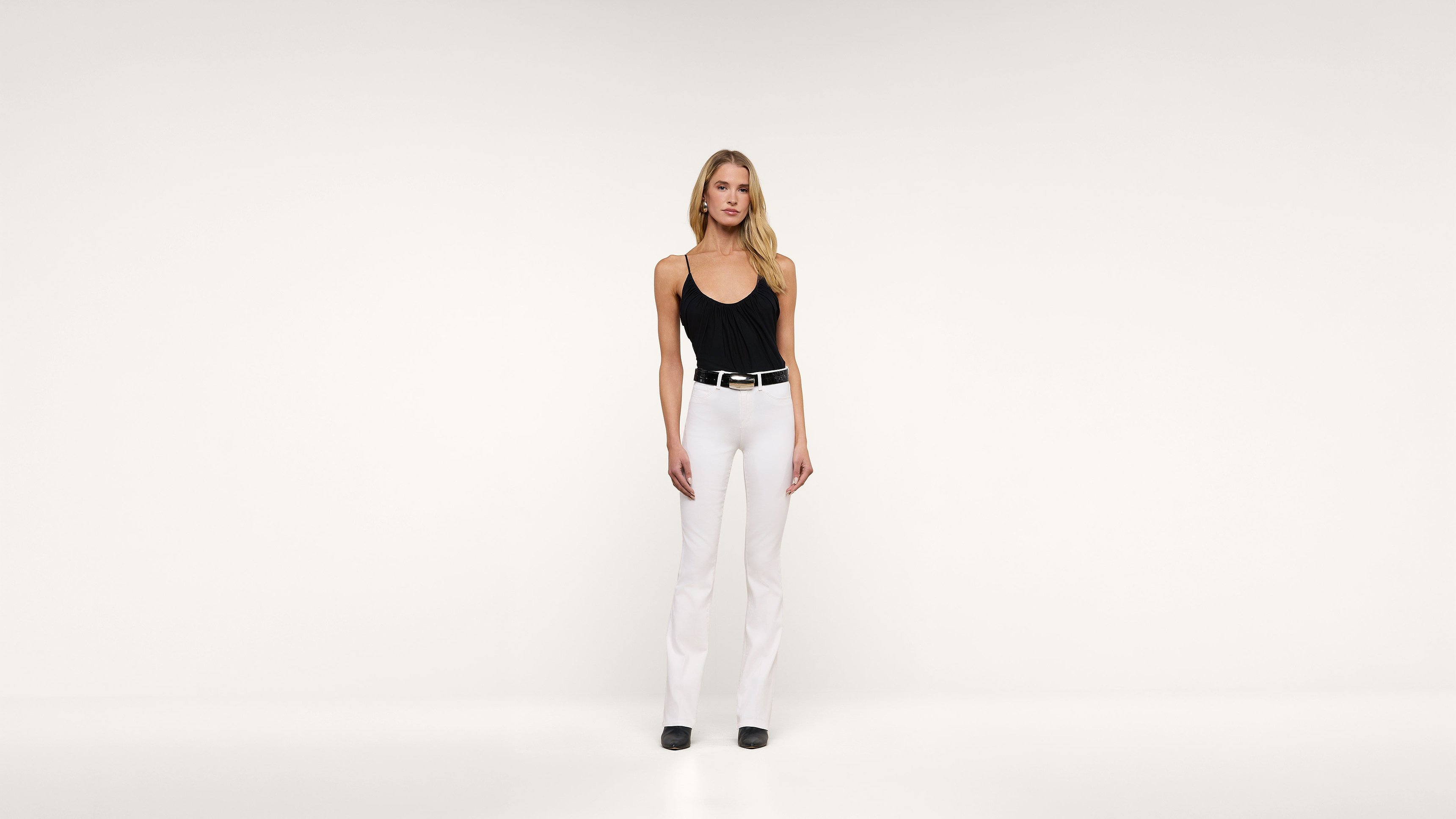 A woman stands against a plain white background wearing a black tank top, white pants, a black belt, and black shoes. She has blonde hair and a neutral expression, with her arms relaxed by her sides.