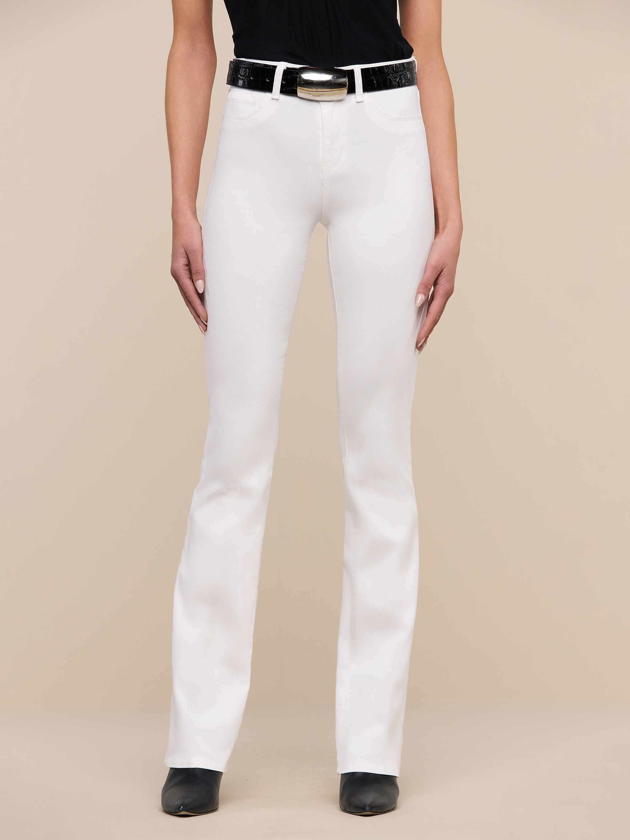 A person stands facing forward, wearing Selma Bootcut Jean in white stretch denim with a contoured waistband, paired with a black belt, black top, and black shoes. The background is plain white, and the person’s face is not visible.