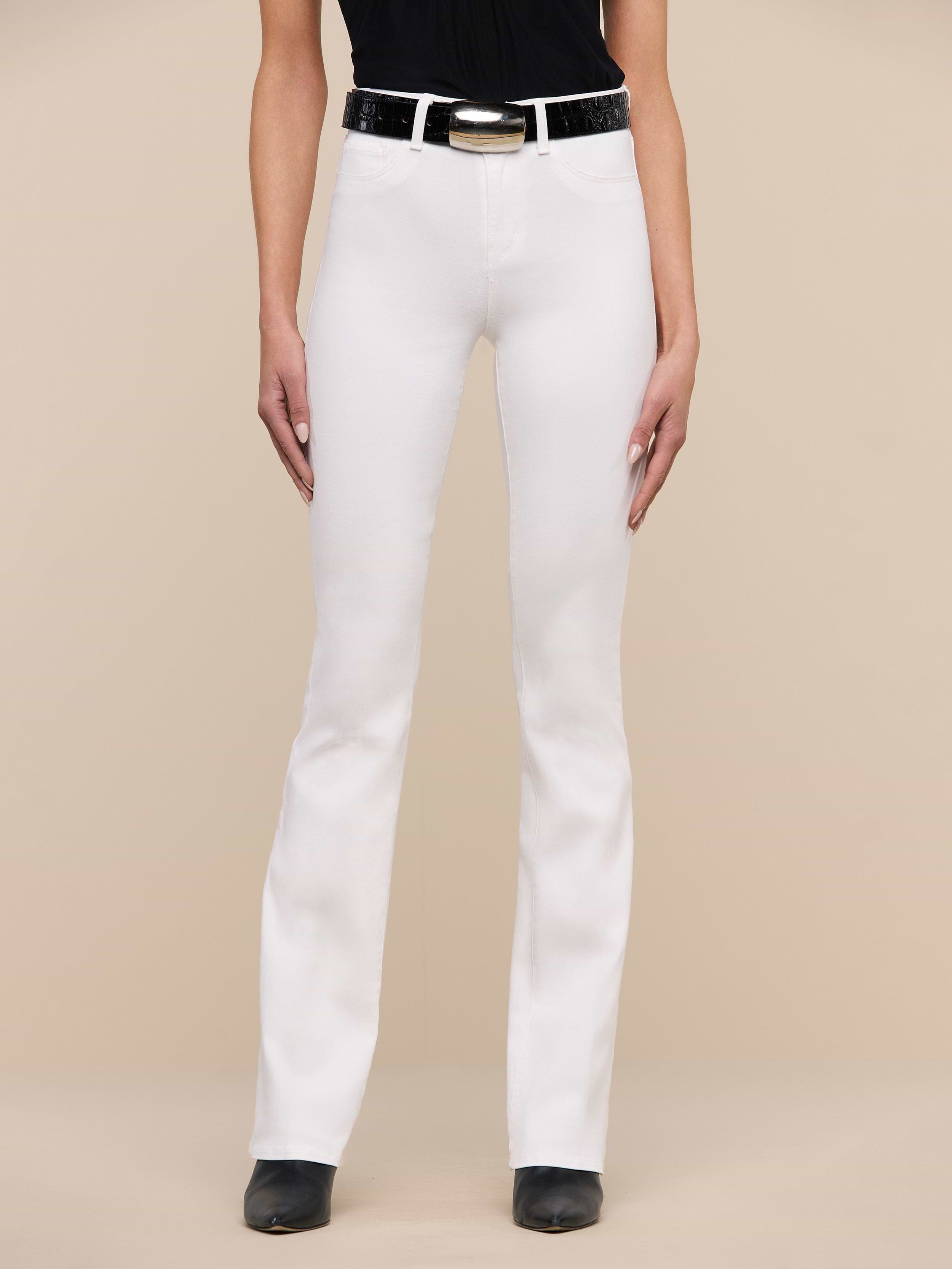 A person stands facing forward, wearing Selma Bootcut Jean in white stretch denim with a contoured waistband, paired with a black belt, black top, and black shoes. The background is plain white, and the person’s face is not visible.