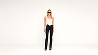A woman wearing sunglasses, a white tank top, black pants, and black heels stands against a plain white background.