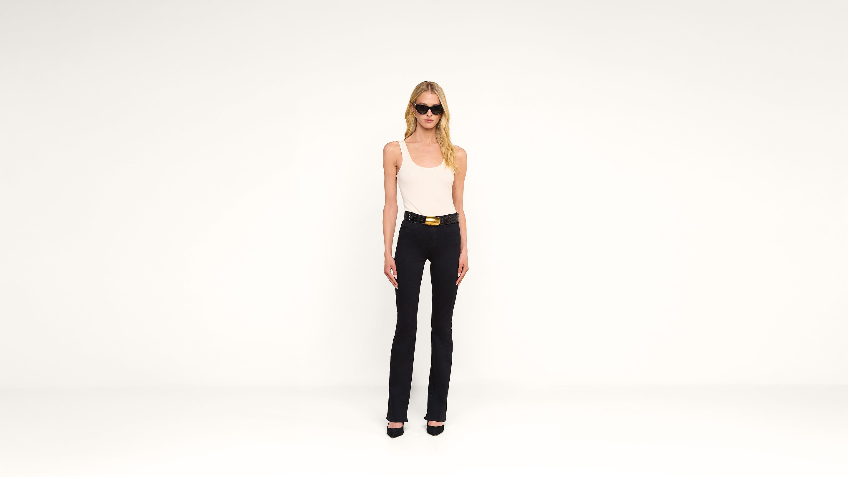 A woman wearing sunglasses, a white tank top, black pants, and black heels stands against a plain white background.