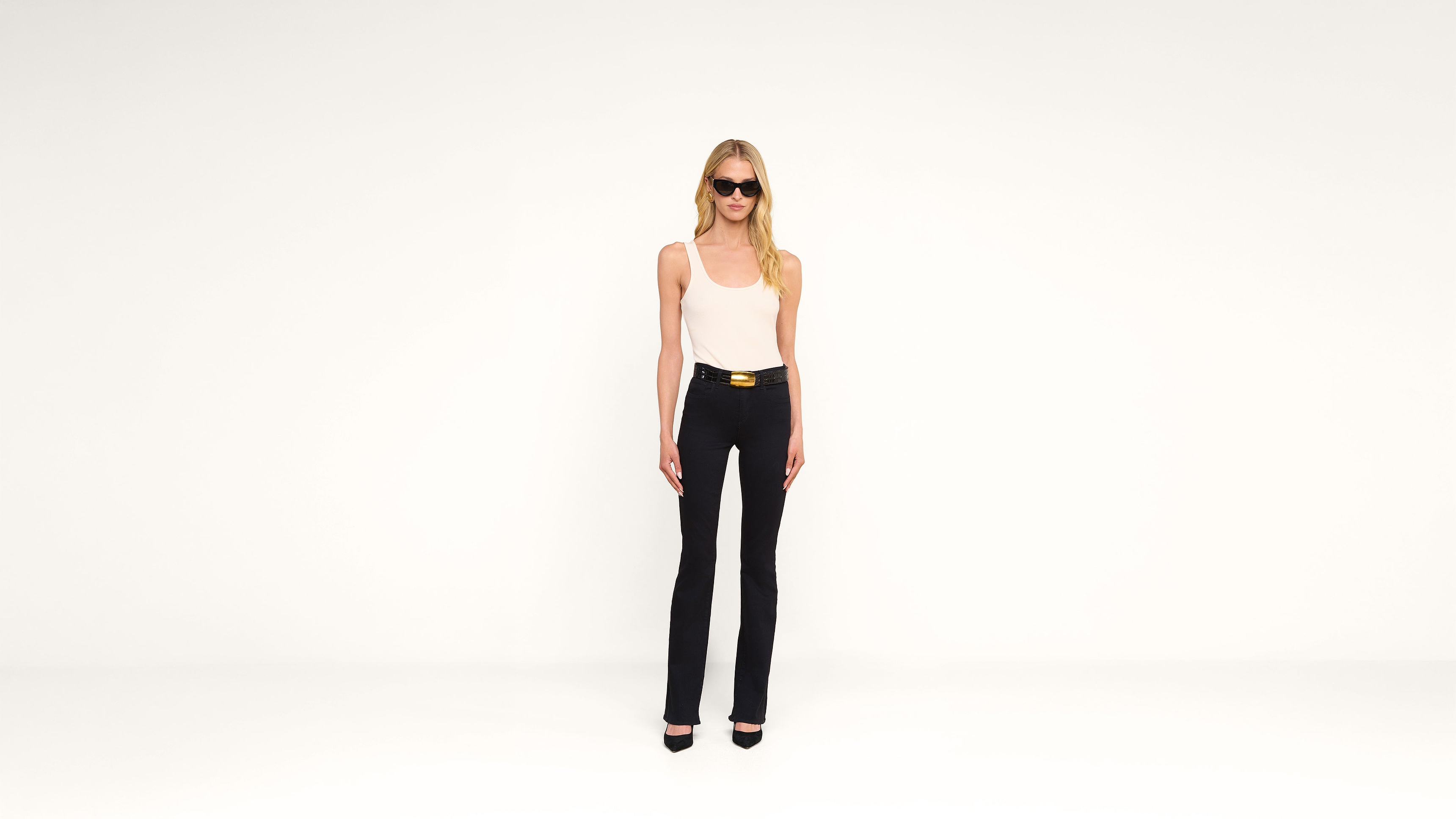 A woman wearing sunglasses, a white tank top, black pants, and black heels stands against a plain white background.