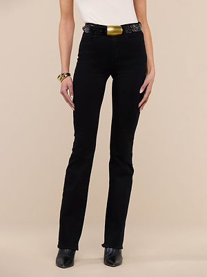 Wearing the Selma Bootcut Jean in true black, a white top, black heeled boots, a black belt with a large gold buckle, and gold bangles, a person stands against a beige background. Their face is not visible.