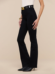Wearing the Selma Bootcut Jean in true black with a high-rise, super stretch fit, a sleeveless white top, a black belt featuring a large gold buckle, and black pointed heels, a person stands against a plain white background.