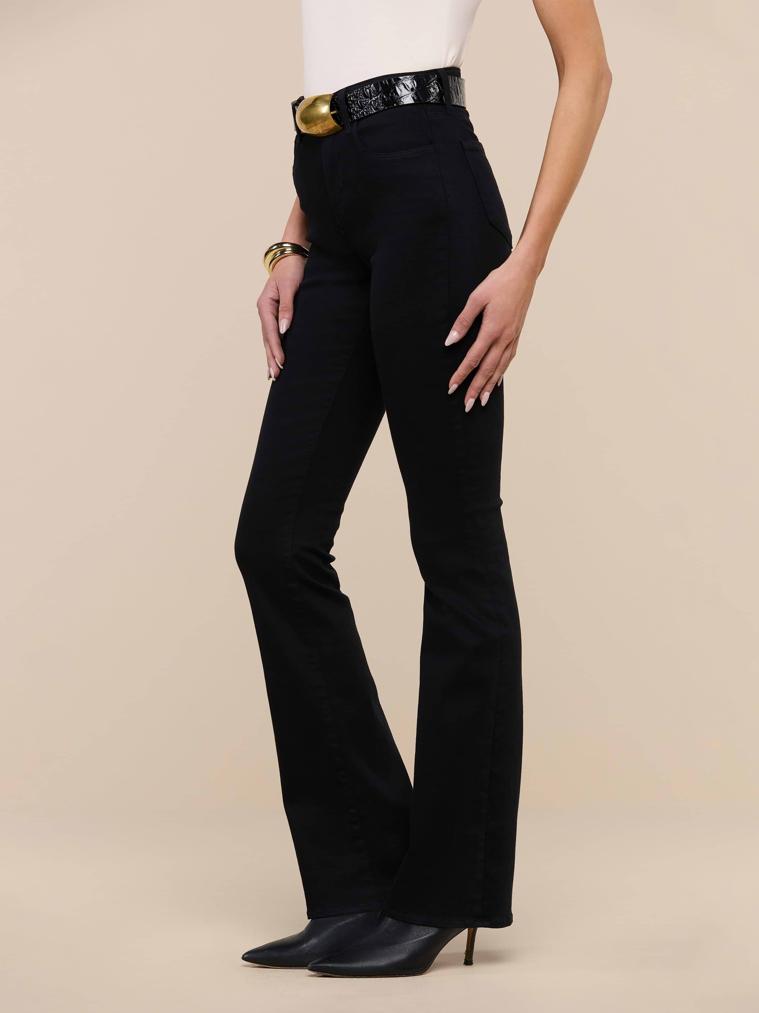Wearing the Selma Bootcut Jean in true black with a high-rise, super stretch fit, a sleeveless white top, a black belt featuring a large gold buckle, and black pointed heels, a person stands against a plain white background.