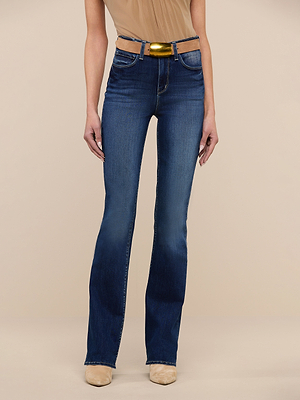 A person wearing the Selma Bootcut Jean in dark blue premium stretch denim with a gold metallic belt, paired with a beige sleeveless top and beige shoes, stands against a plain beige background.