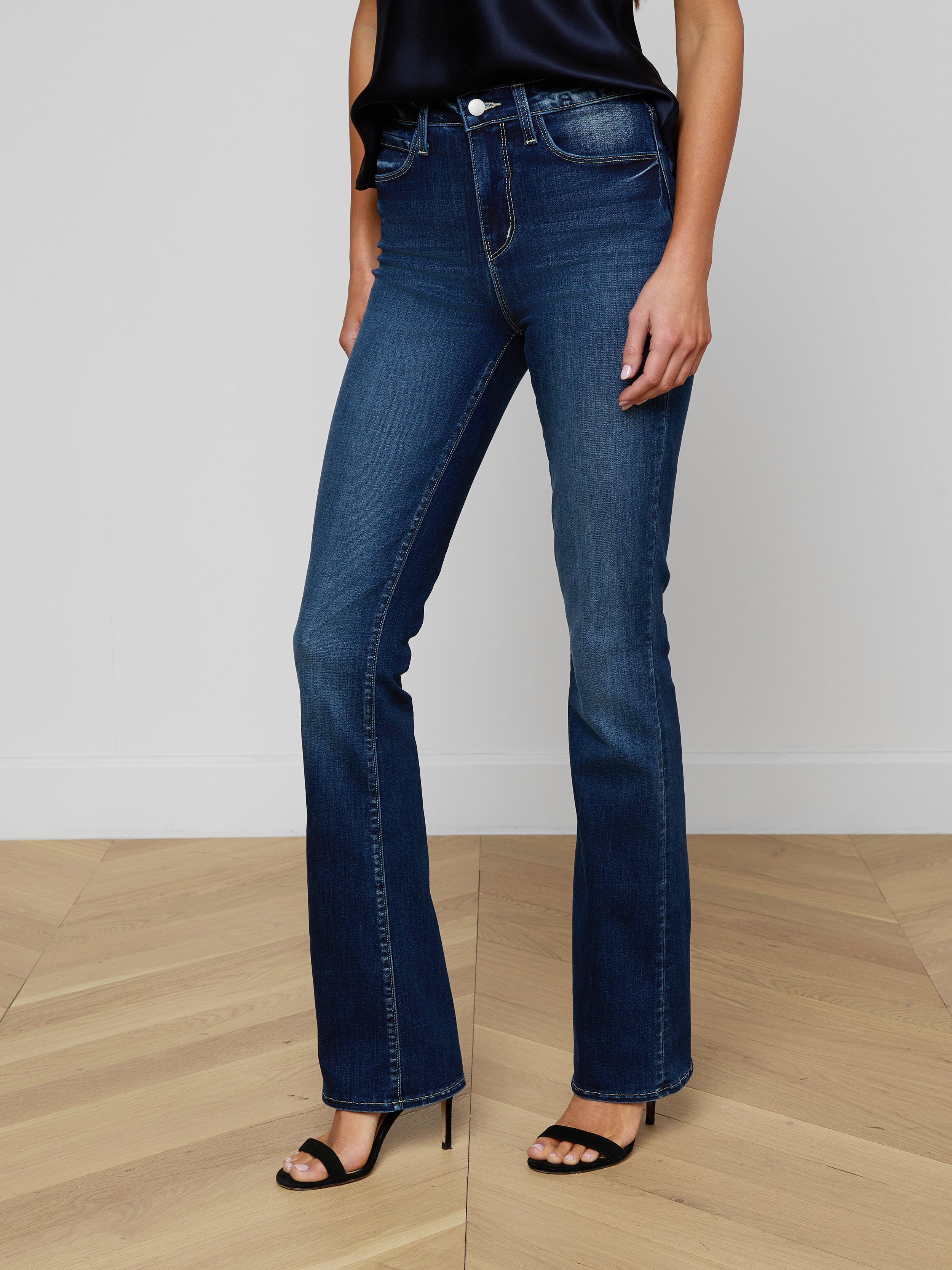 Wearing the Selma Bootcut Jean in fitted blue stretch denim, a brown belt, a beige sleeveless top, and tan pointed-toe heels, the person stands against a plain white background.