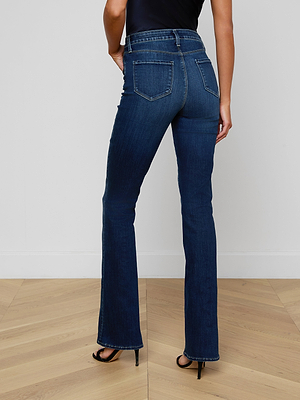A woman stands indoors on a wooden floor, facing away from the camera, wearing Selma Bootcut Jeans in dark blue, paired with a black sleeveless top and black high-heeled sandals.