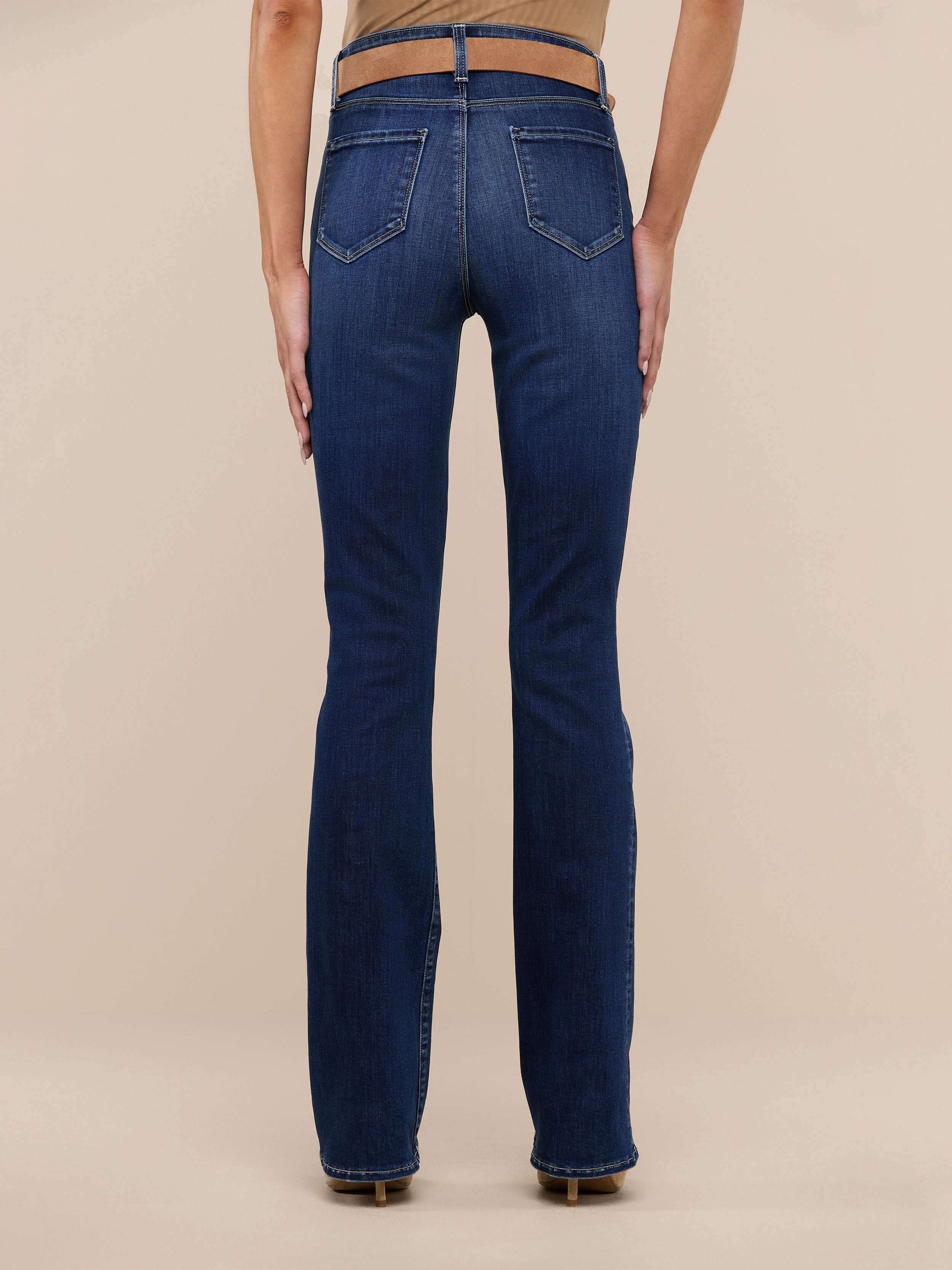 A person is standing facing away, wearing a beige sleeveless top, beige heels, and the Selma Bootcut Jean—a pair of dark blue high-rise slim bootcut jeans crafted from stretch denim—against a plain white background.