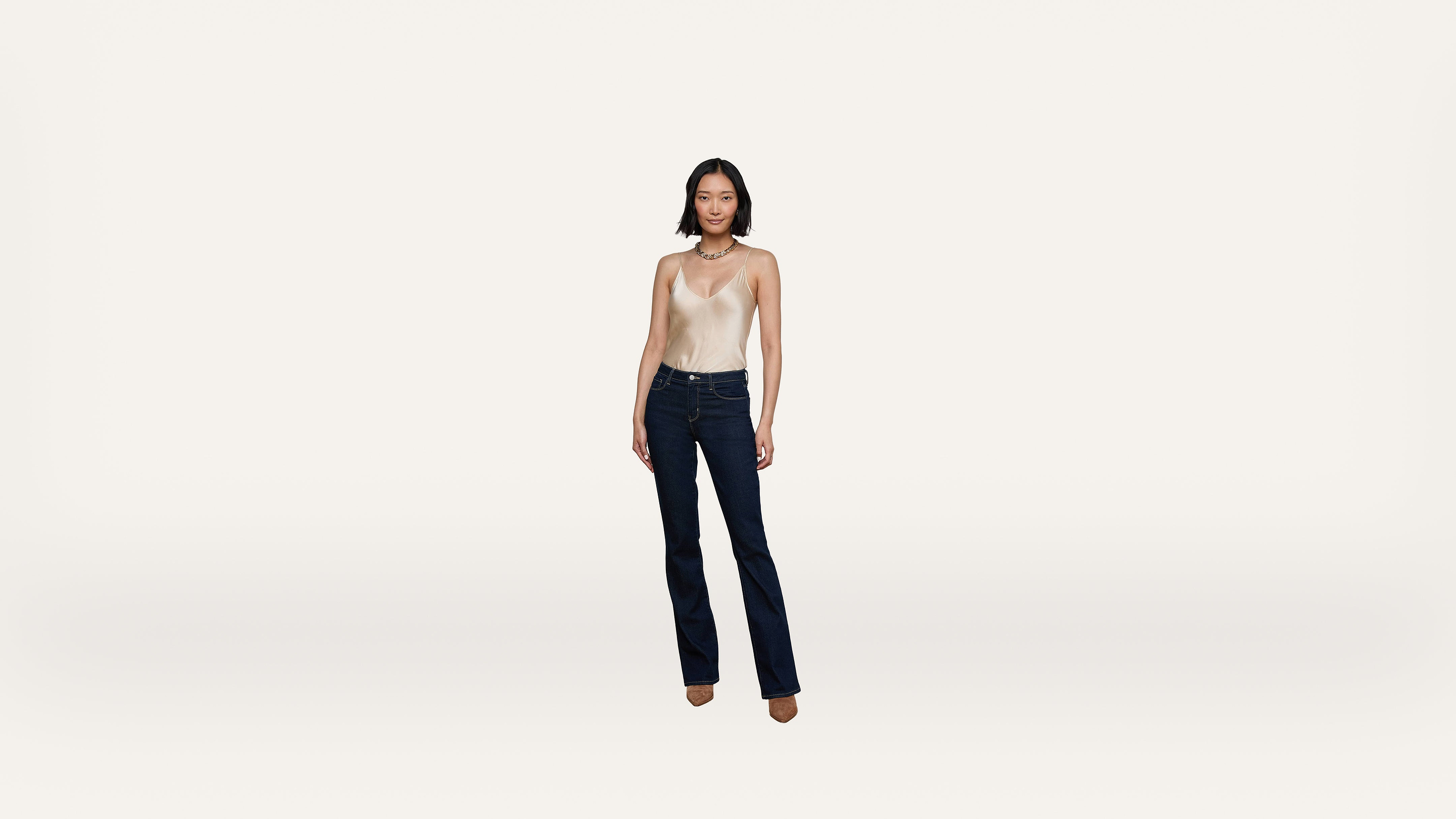 A woman stands facing forward, wearing a white sleeveless top, blue jeans, and brown shoes, against a plain white background.
