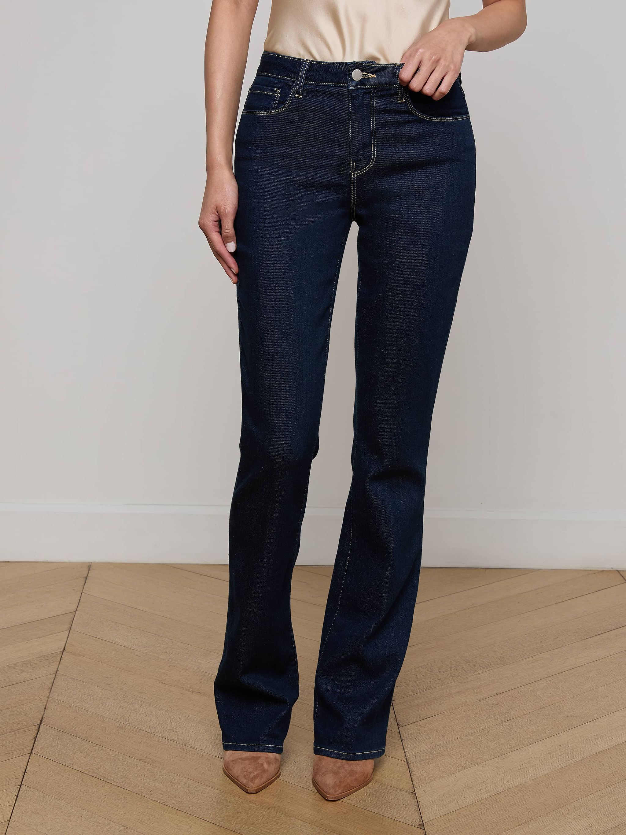 A person wearing the Selma Bootcut Jean in high-rise dark blue stretch denim and tan pointed-toe shoes stands against a plain light background, with their face not visible.
