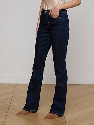 A person wearing L'AGENCE Selma Bootcut Jeans in dark blue, paired with a beige top and brown pointed-toe shoes, stands on a light wooden floor against a plain white wall.