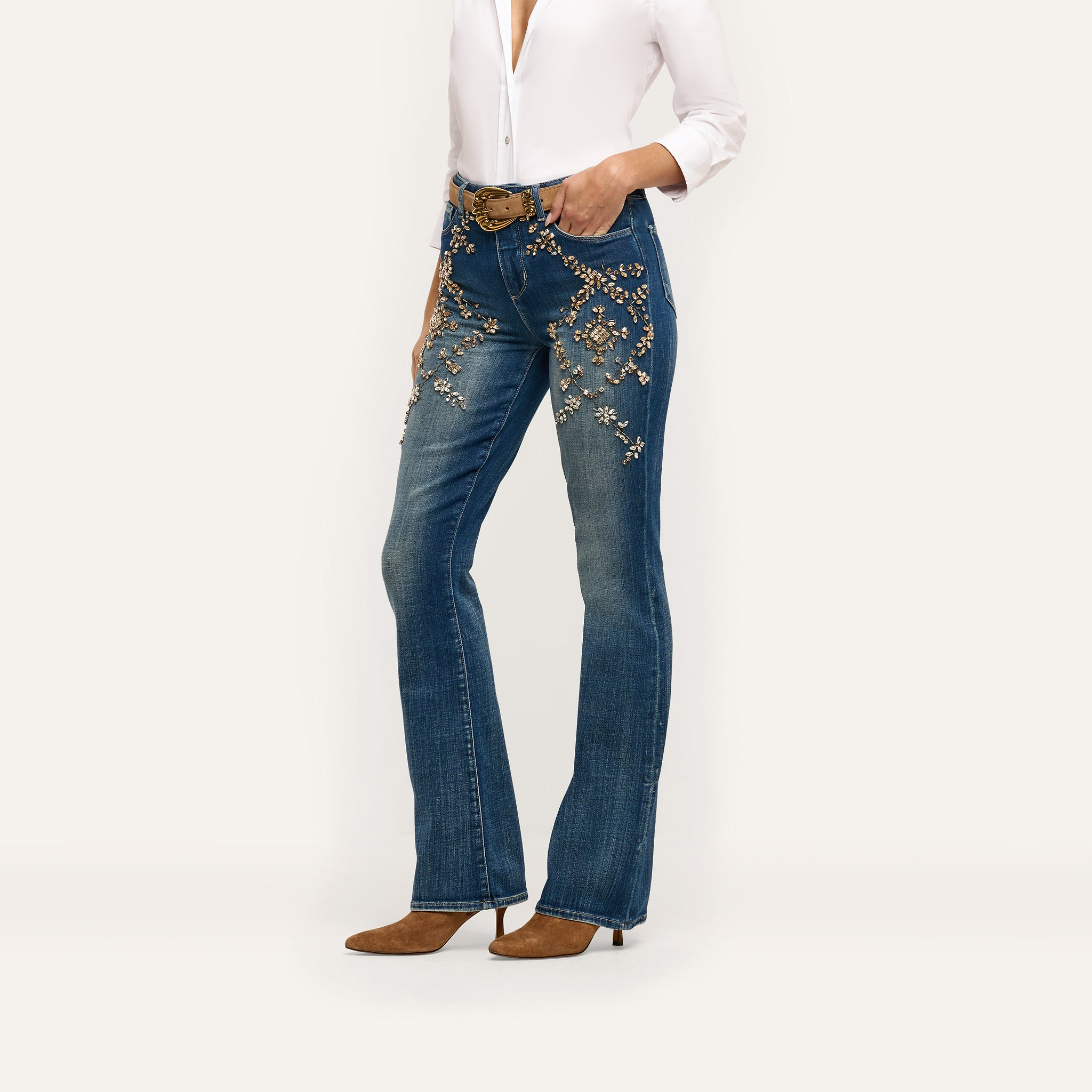 Selma Embellished Bootcut Jean in blue, featuring gold-toned floral embellishments and crystal details on the hips, paired with a white button-down shirt and brown pointed-toe heels.
