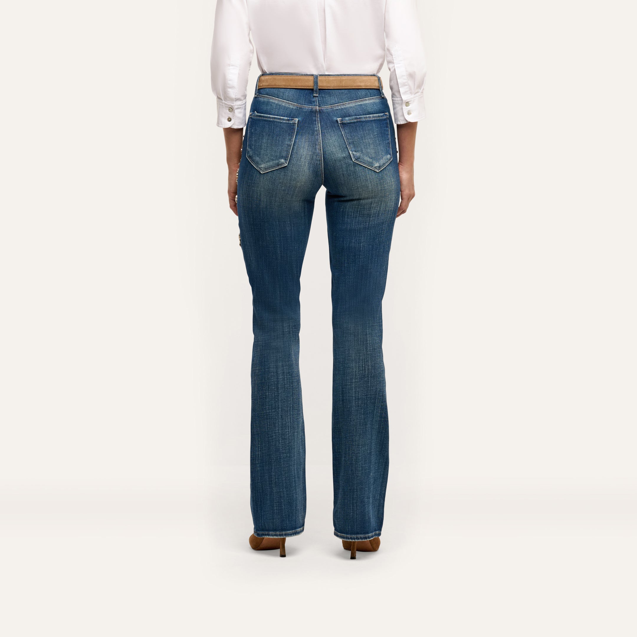 Rear view of blue Selma Embellished Bootcut Jean with crystal-embellished pockets, worn with a white button-down shirt and a thin tan belt.