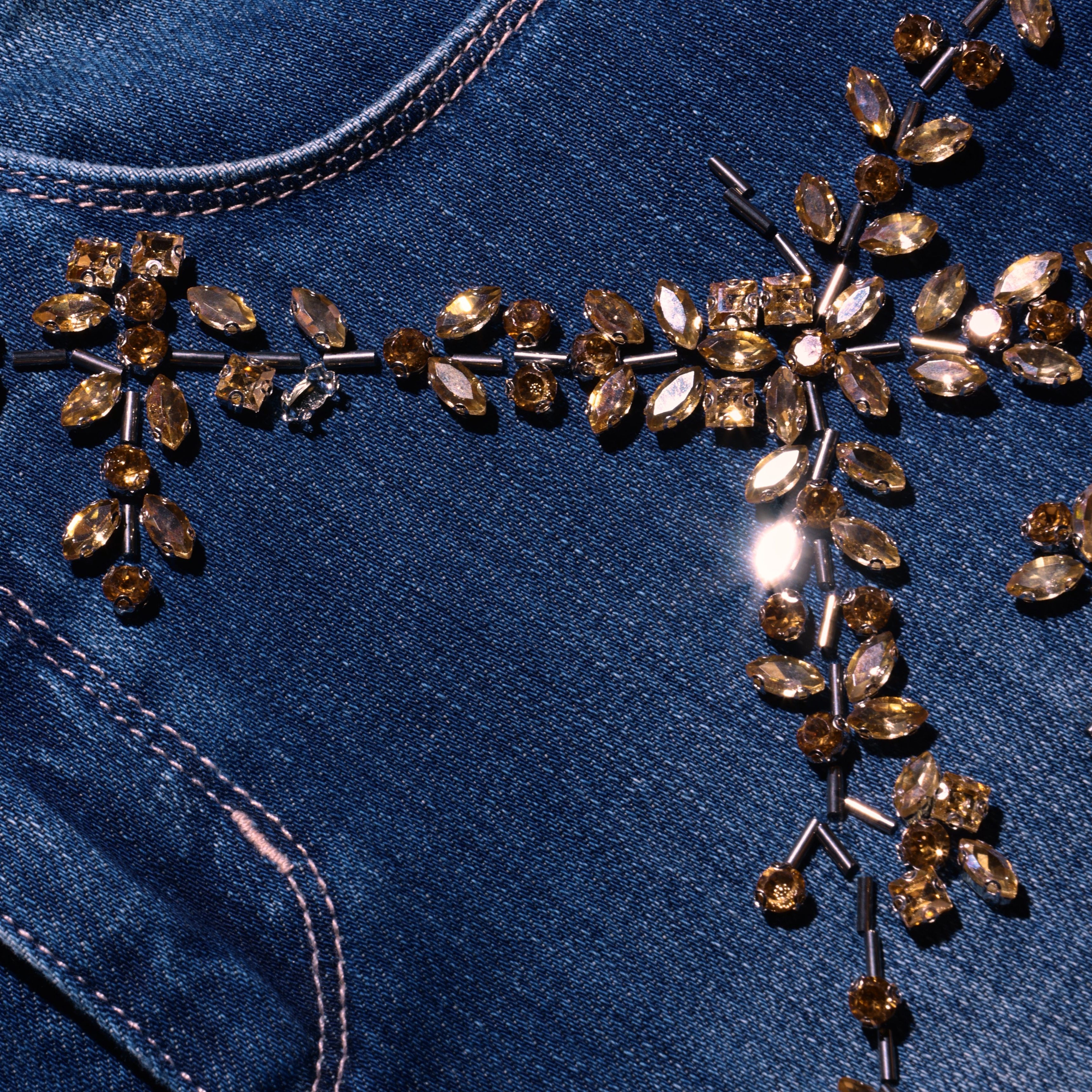 Close-up of blue denim Selma Embellished Bootcut Jean, featuring a floral pattern of amber-colored crystals and metal beads.