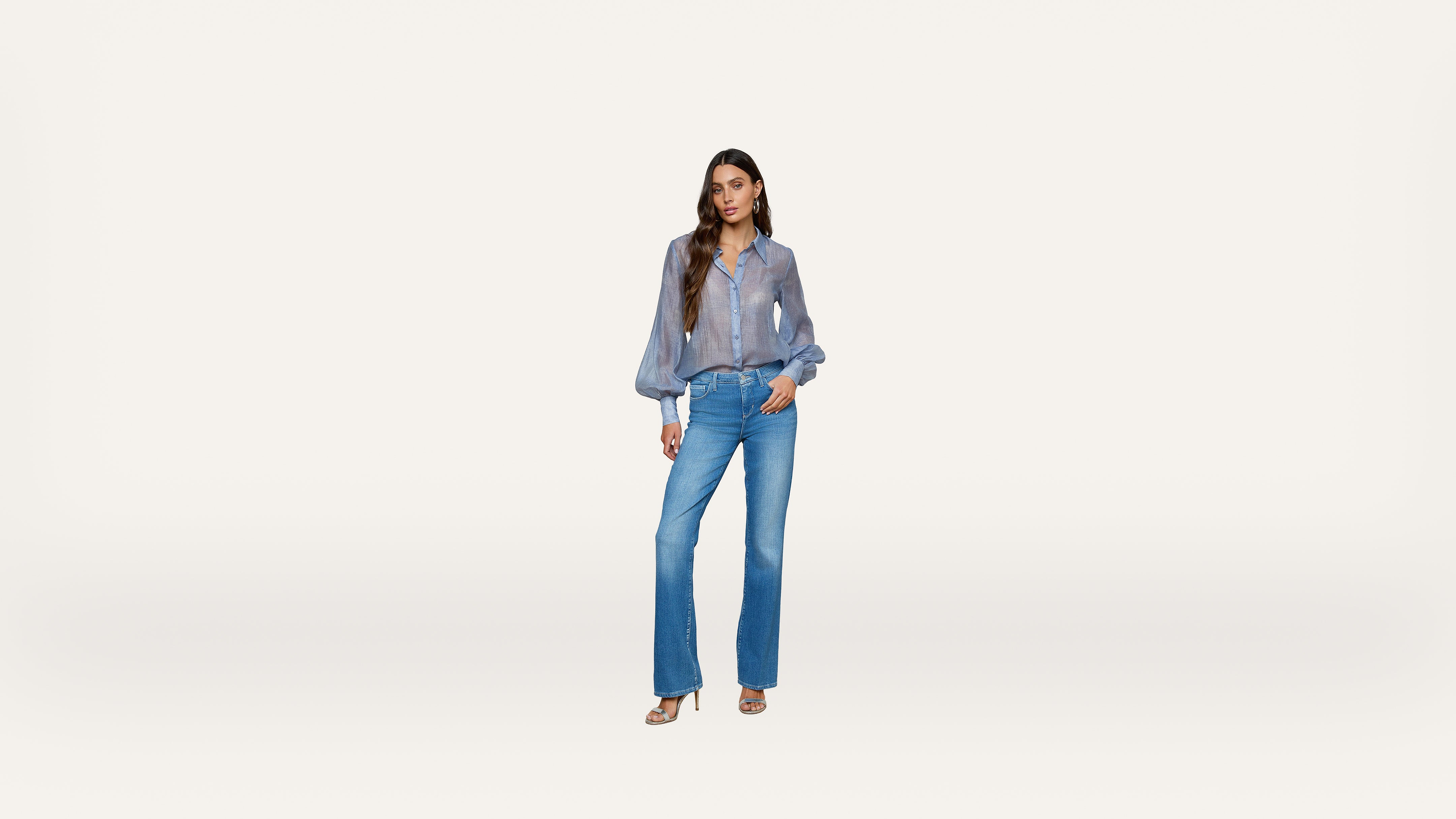 Woman standing against a white background, wearing a sheer long-sleeve blue blouse, high-waisted flare blue jeans, and beige heels, with her left hand in her pocket and her hair styled in loose waves.