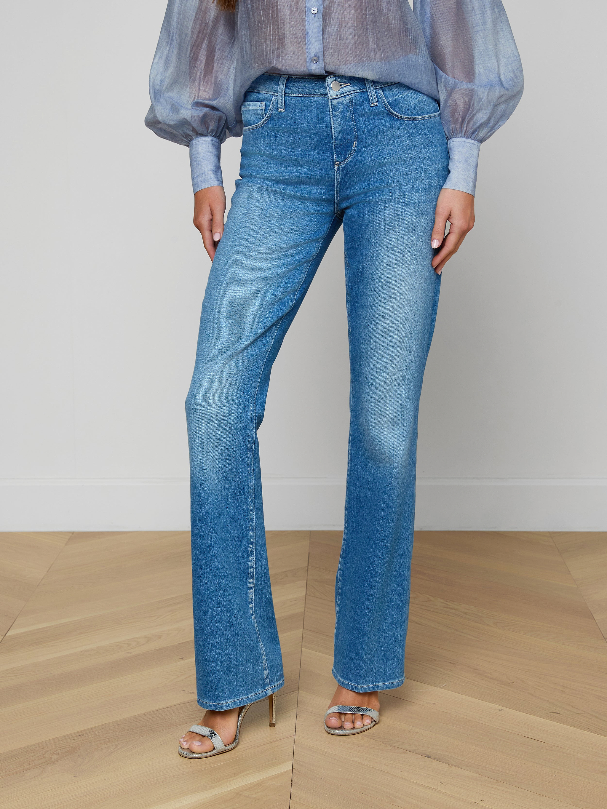 A person wearing the Selma Bootcut Jean in light blue stretch denim with a contoured waistband, paired with a sheer blue blouse with puffy sleeves and beige heeled sandals, standing against a plain background.