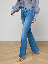 A woman wearing the Selma Bootcut Jean in light blue stretch denim, paired with a sheer blue long-sleeve blouse and silver high-heeled sandals, walks against a plain background.