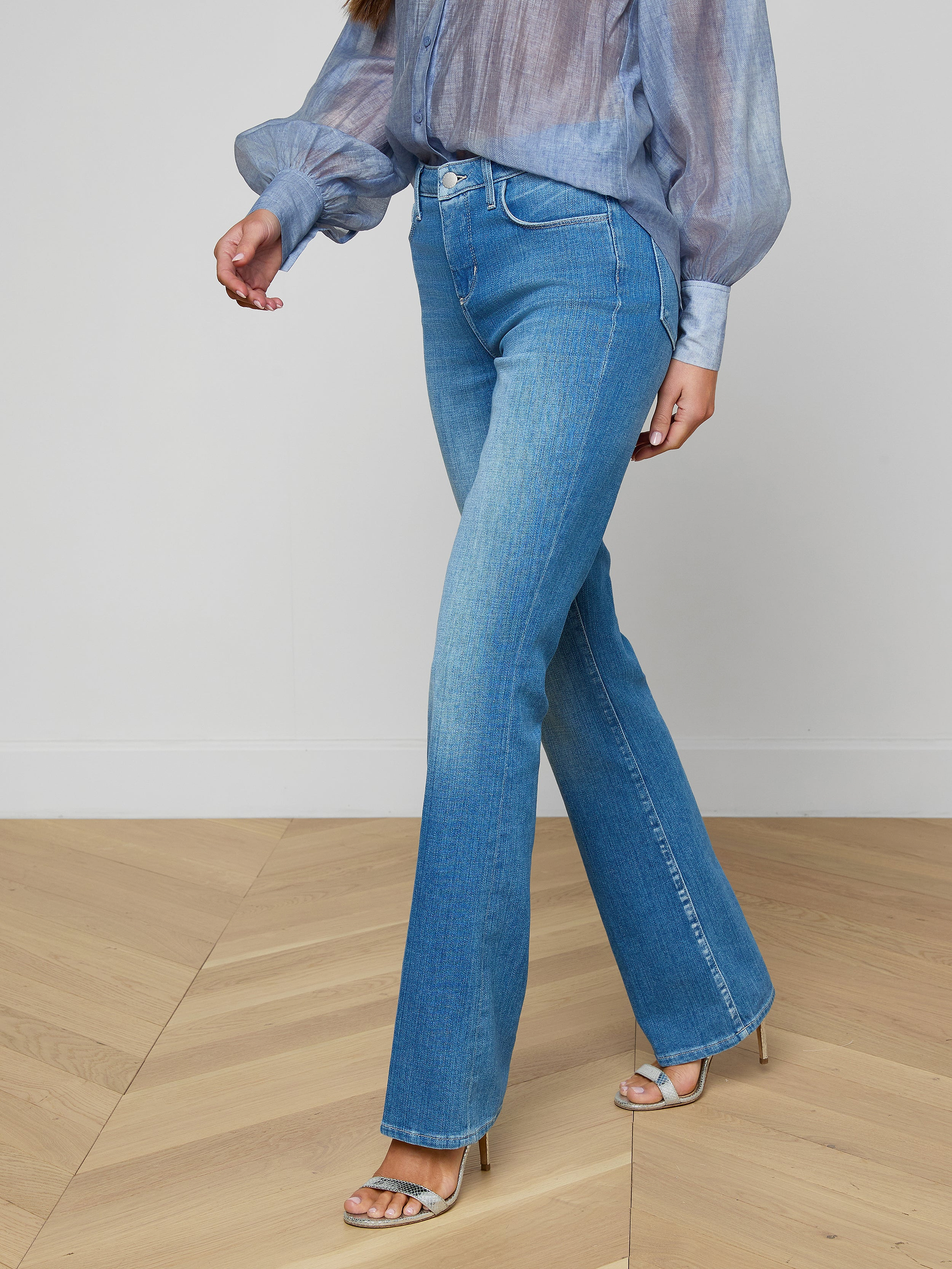 A woman wearing the Selma Bootcut Jean in light blue stretch denim, paired with a sheer blue long-sleeve blouse and silver high-heeled sandals, walks against a plain background.