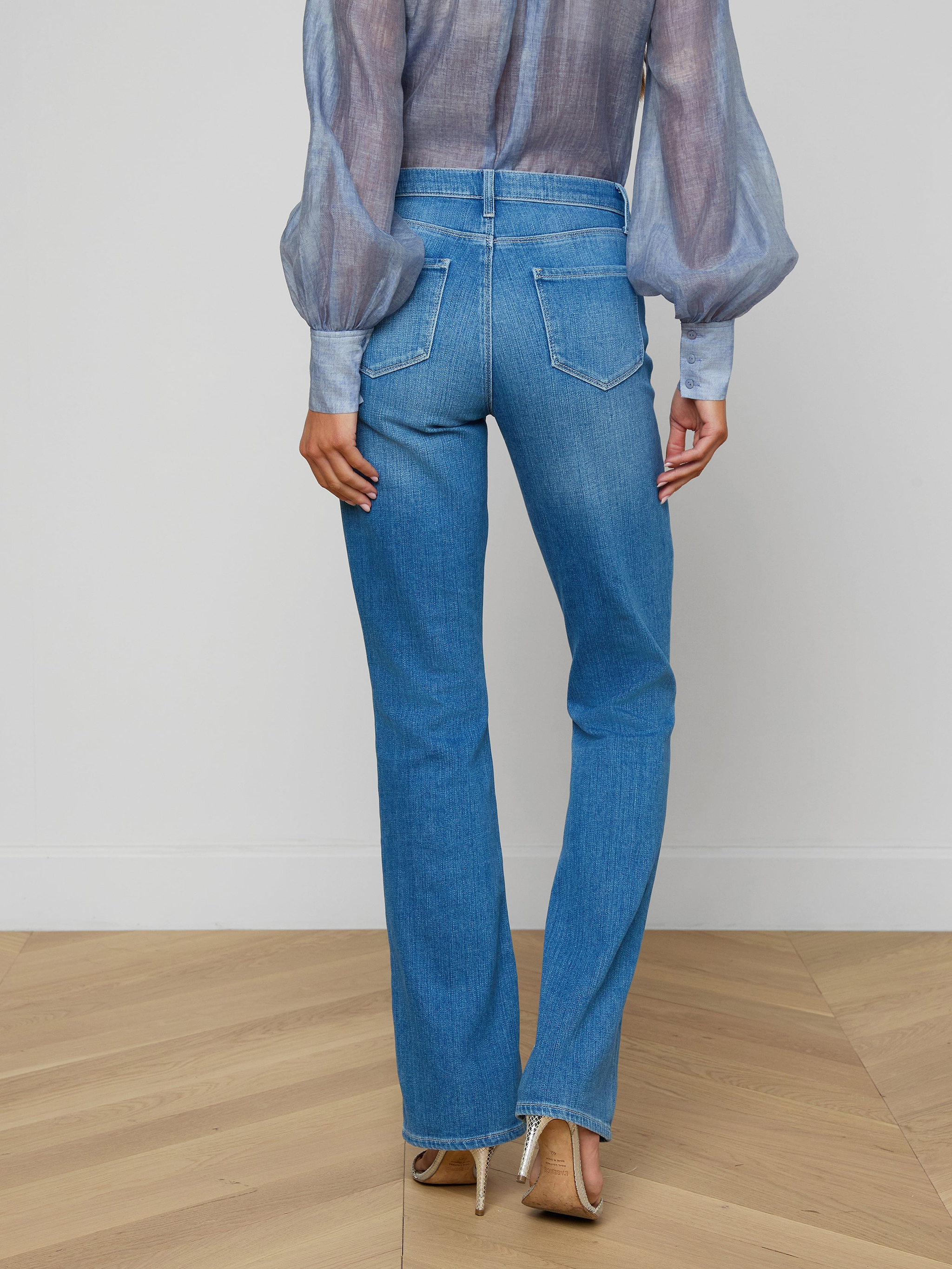 A person stands facing away against a plain, light background, wearing a sheer long-sleeve blouse, the Selma Bootcut Jean in blue featuring a contoured high-rise waistband, and beige heels, showcasing the back of the outfit.