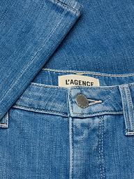 Close-up of the Selma Bootcut Jean in blue denim, showcasing a metal button, contoured waistband, belt loop, and an interior clothing label with the LAGENCE brand name. A denim shirt is partially draped over the jeans.