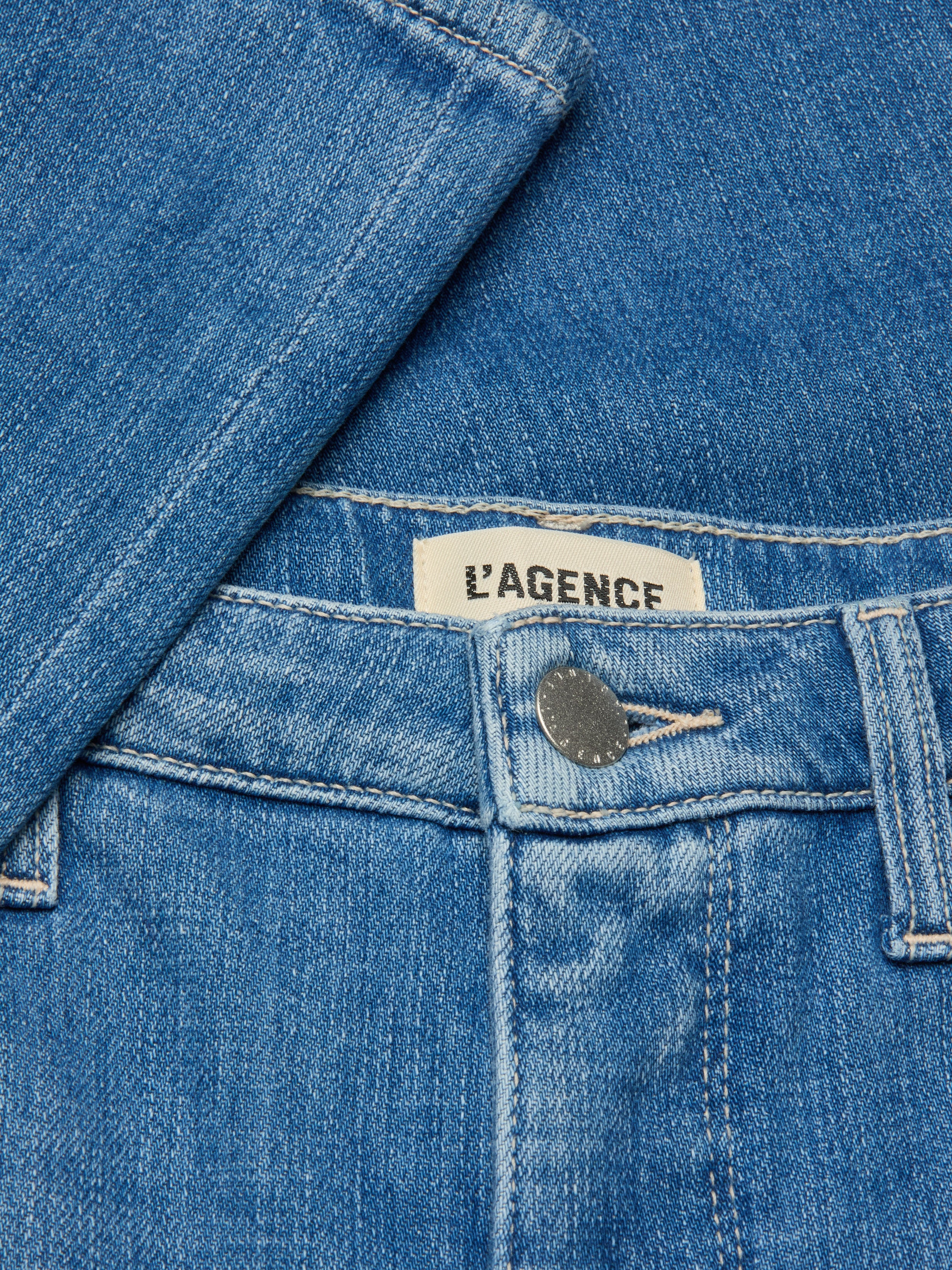 Close-up of the Selma Bootcut Jean in blue denim, showcasing a metal button, contoured waistband, belt loop, and an interior clothing label with the LAGENCE brand name. A denim shirt is partially draped over the jeans.