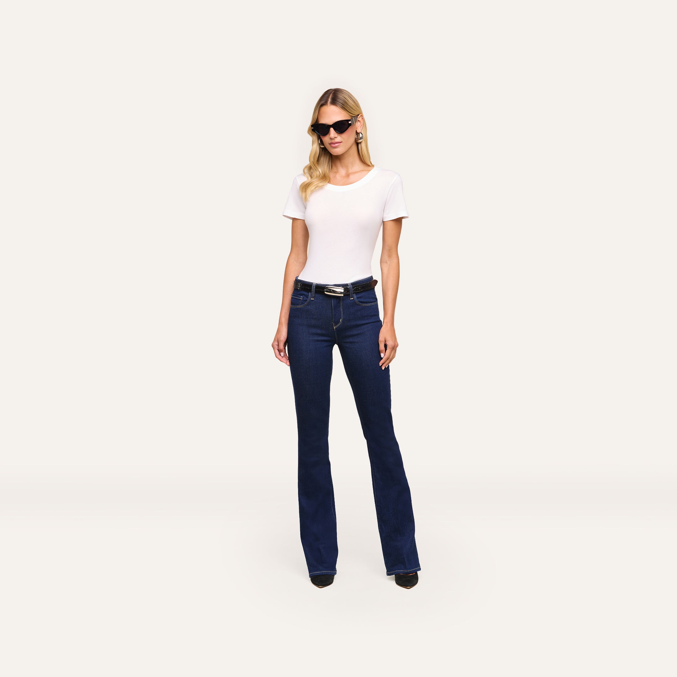 A woman in sunglasses, a white short-sleeve top, and Selma Bootcut Jean in high-rise dark blue stretch denim stands against a plain light background.