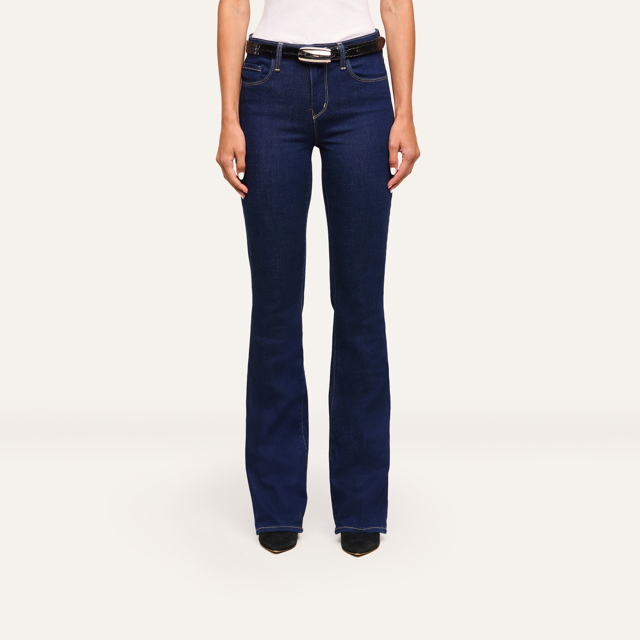 A person wears the Selma Bootcut Jean in dark blue stretch denim with a white top and black shoes, standing against a plain white background.