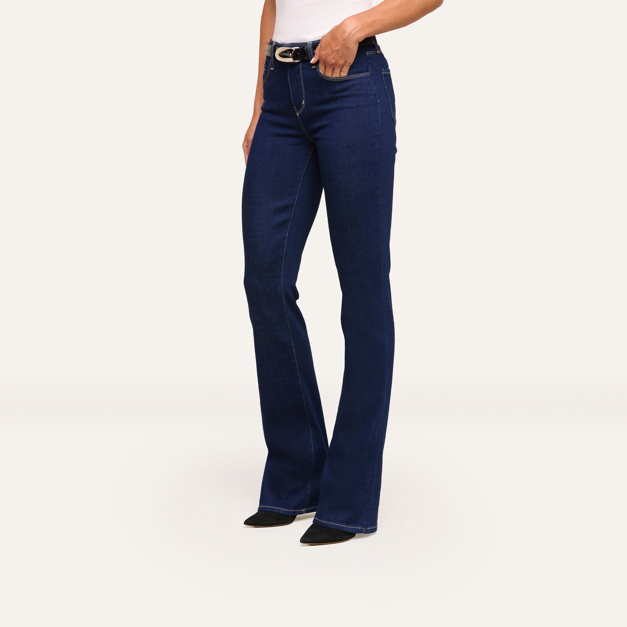 A person wears Selma Bootcut Jean in dark blue stretch denim, paired with a white top and black pointed shoes, standing against a plain light background.